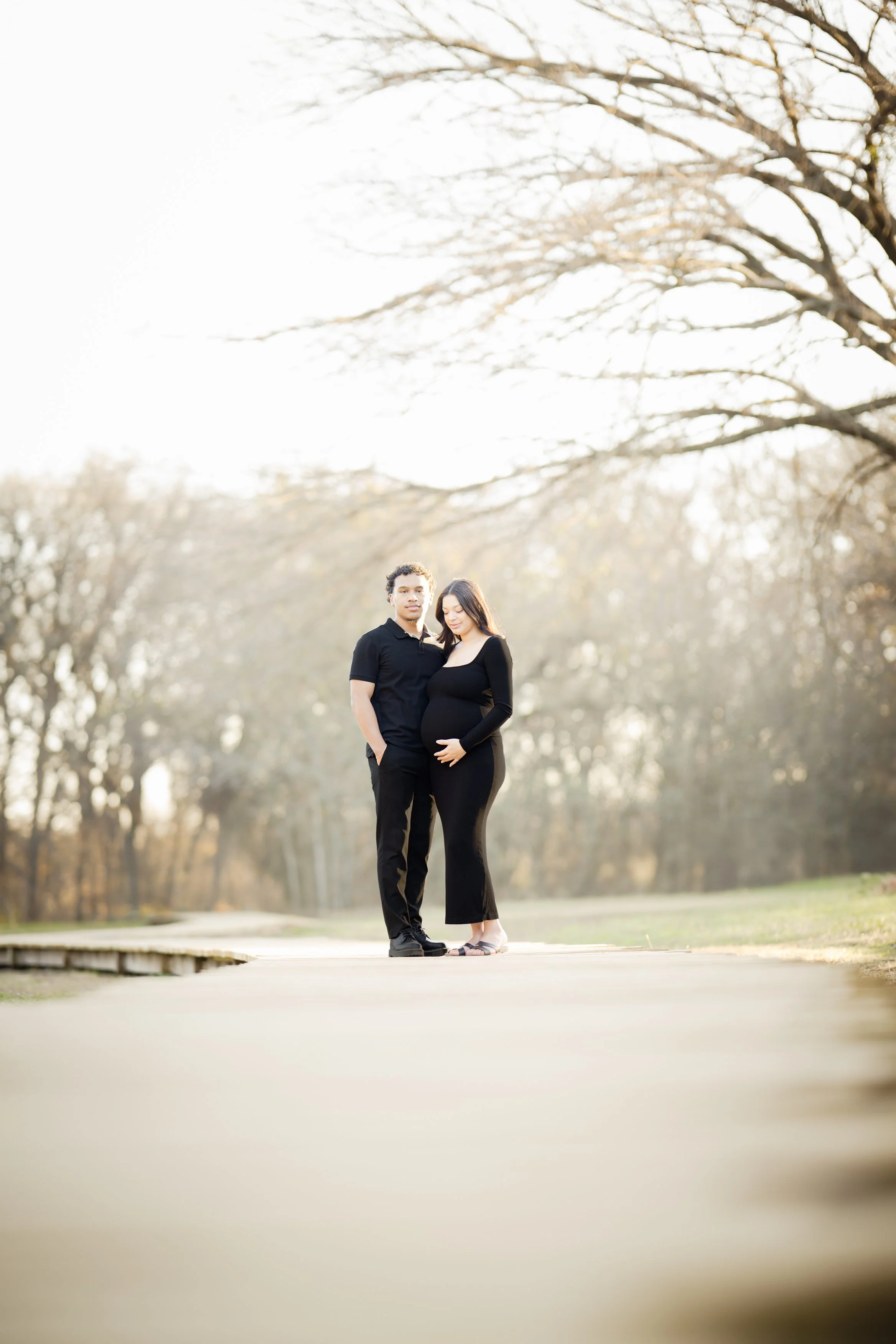 A wide-angle maternity portrait bathed in soft, golden light. The couple stands together on a quiet path framed by trees, evoking serenity, anticipation, and the beauty of a new chapter unfolding.
