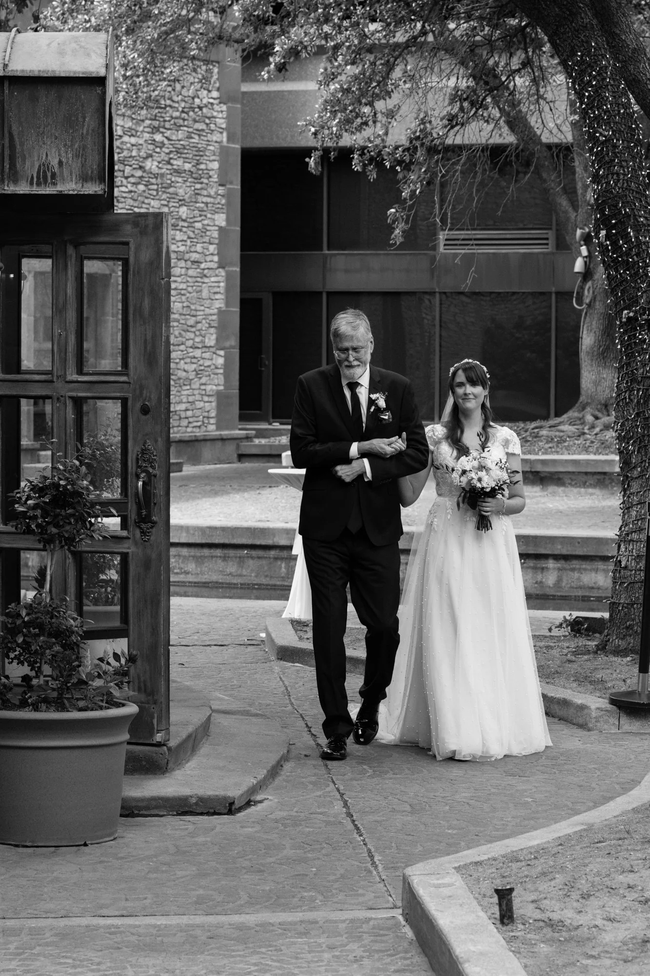 A black-and-white wedding portrait of a bride walking arm-in-arm with her father down a stone pathway, framed by trees and architectural details, capturing a quiet and emotional pre-ceremony moment.