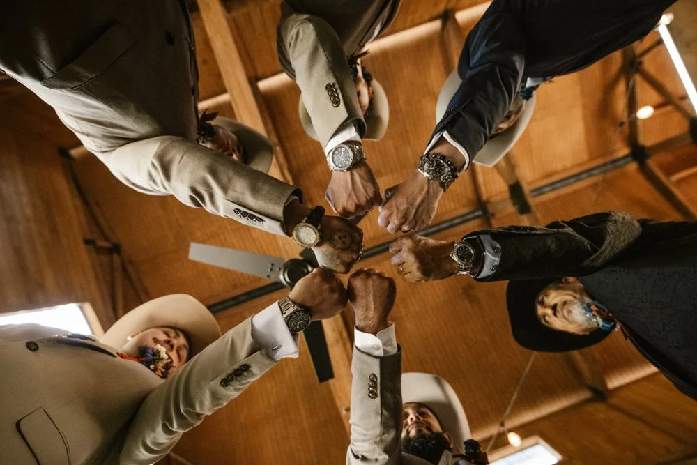 Groom and groomsmen wearing cowboy hats and tan suits joining fists together before a rustic Texas wedding ceremony.