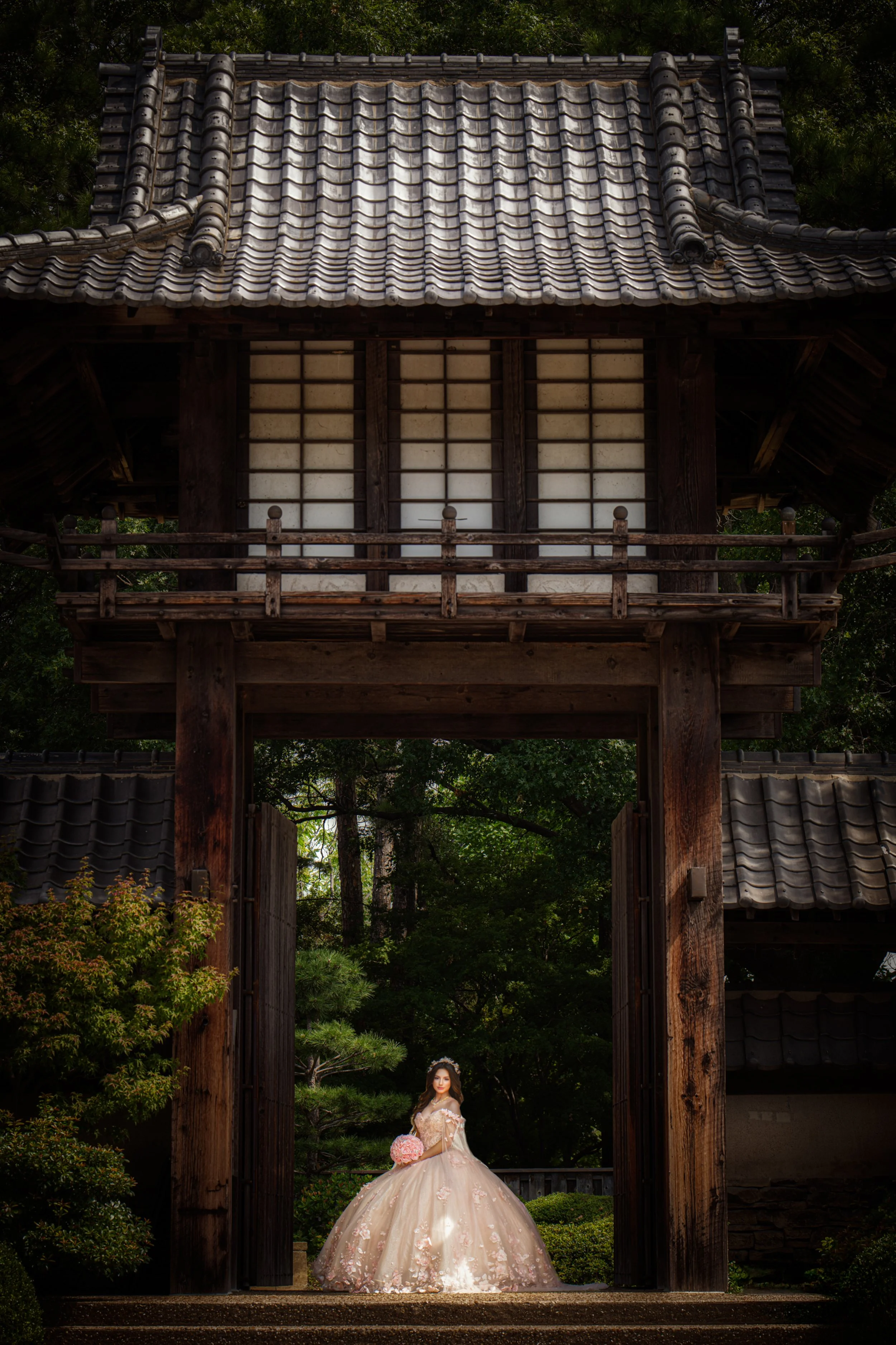 A dramatic wide-angle portrait of the quinceañera framed by a traditional garden gate, emphasizing symmetry, scale, and timeless elegance as she stands confidently in her embellished quinceañera gown.