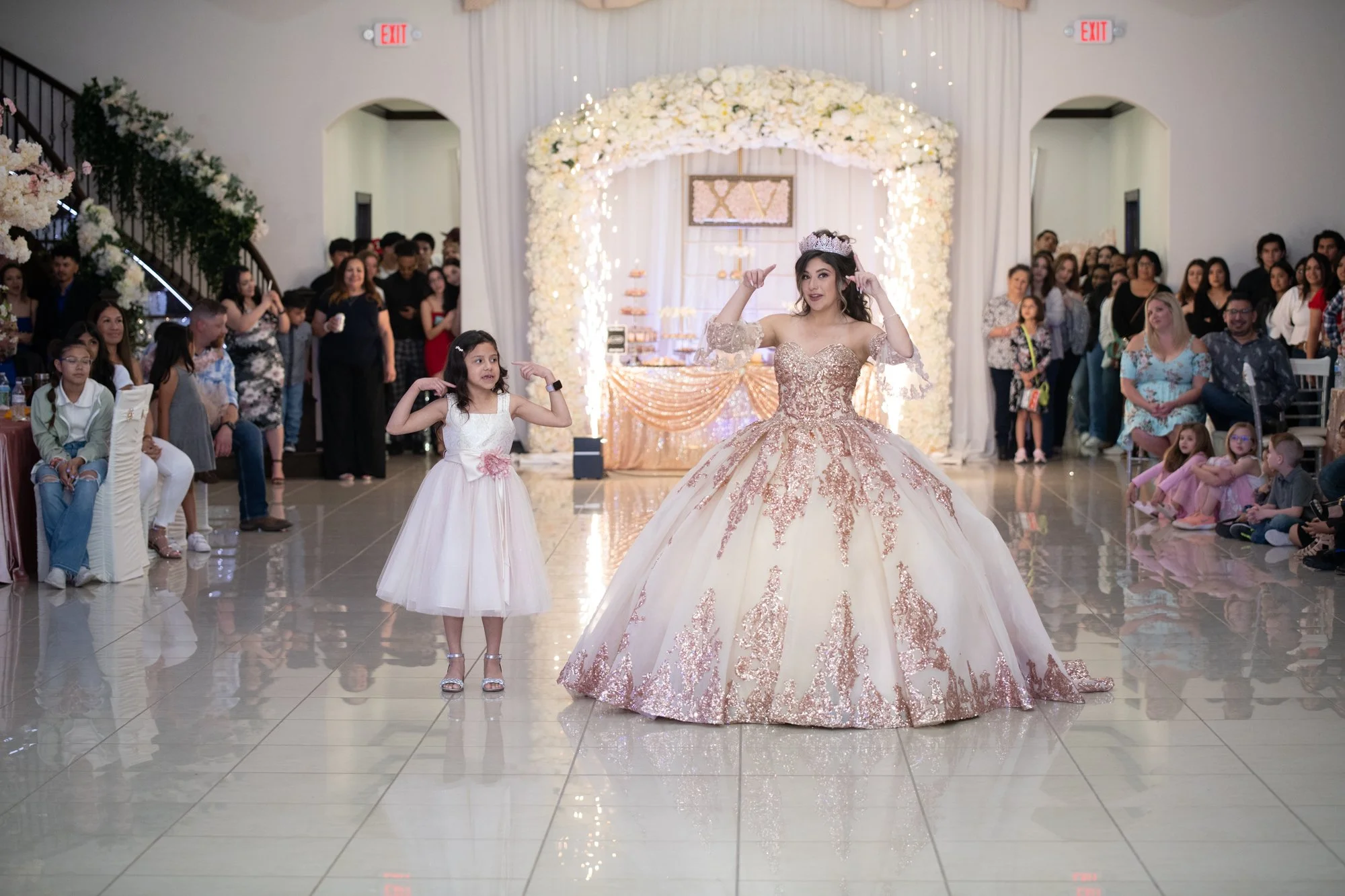 A sweet and playful moment as Katelyn dances alongside a younger family member in front of a floral-lit backdrop. Guests line the walls watching proudly as the spotlight centers on sisterhood, confidence, and joy.