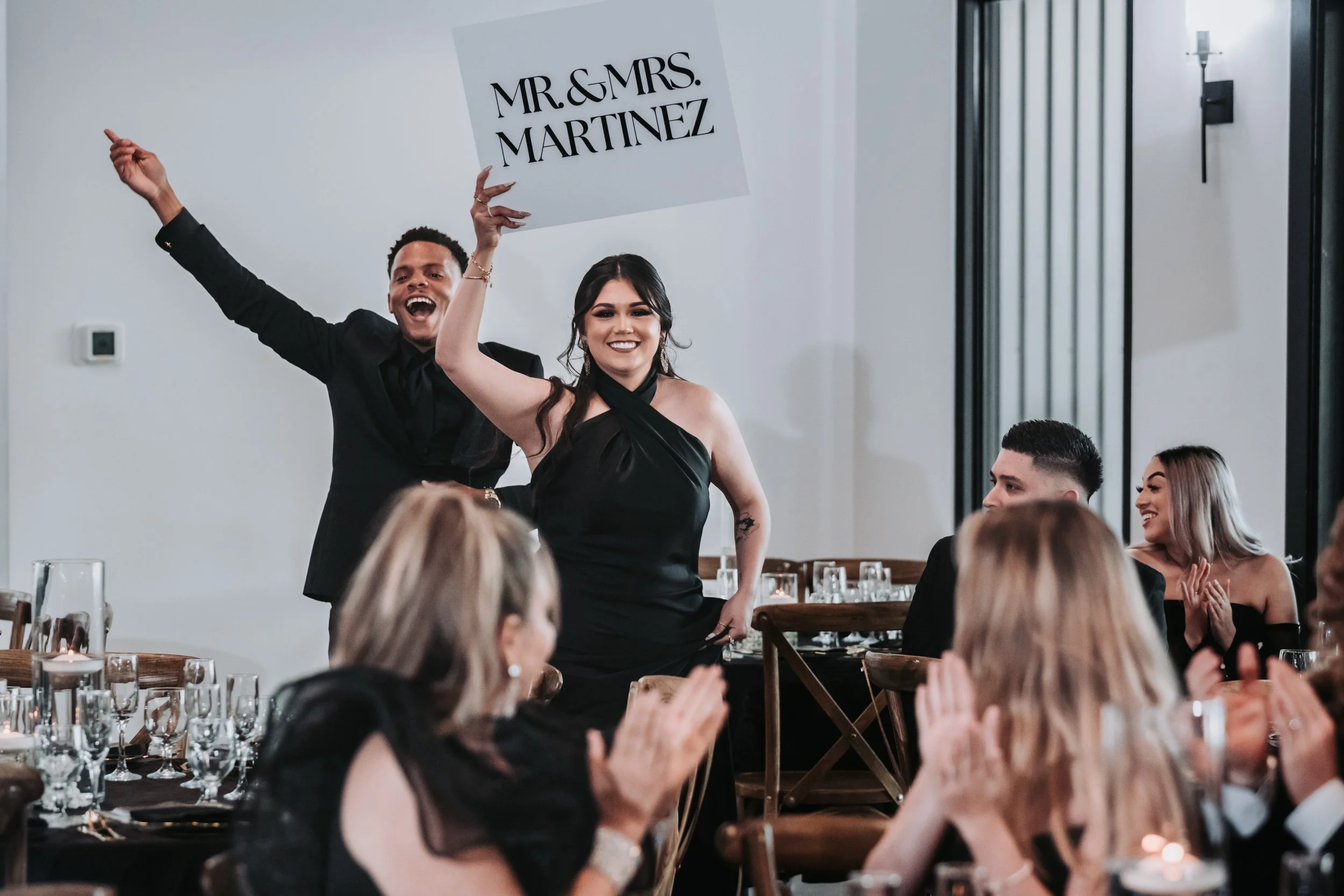 The couple makes their reception entrance holding a “Mr. & Mrs.” sign as guests cheer, celebrating the beginning of their marriage with energy and joy.