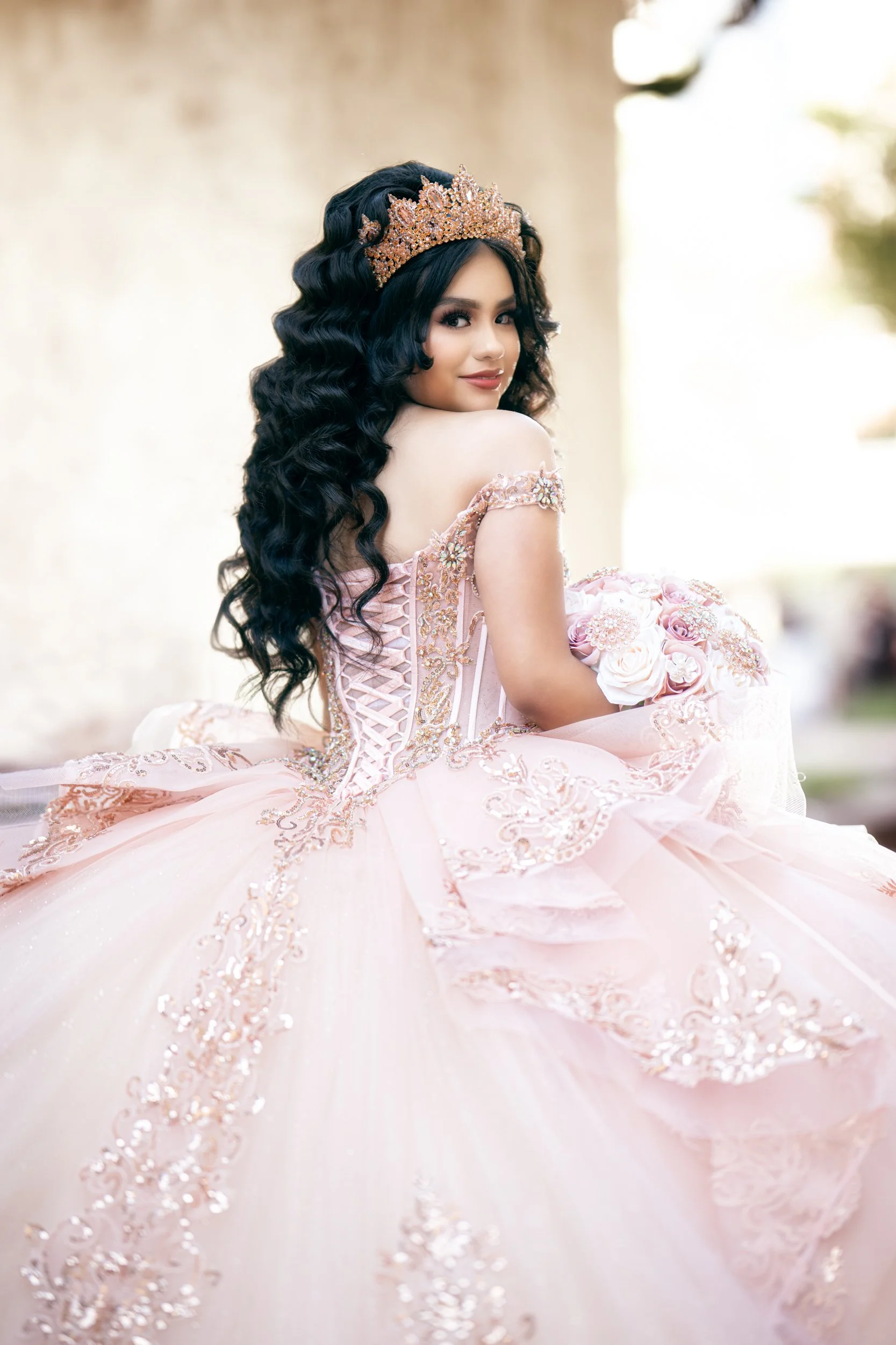 A close-up over-the-shoulder portrait highlights the quinceañera’s soft expression, detailed crown, and delicate embroidery on her dress. The natural light creates a warm and dreamy atmosphere.