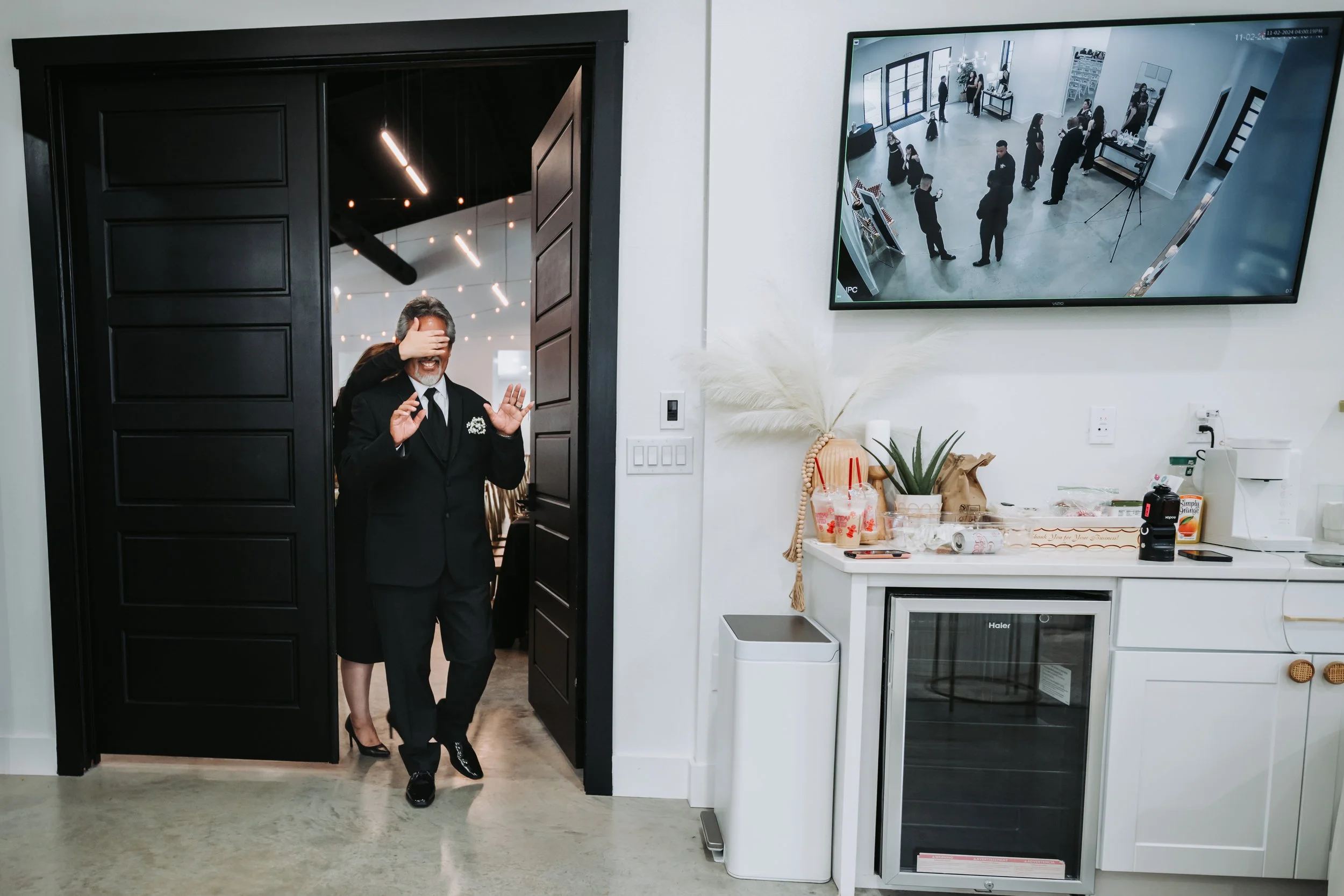 The father enters the room for his first look, hands raised in surprise and emotion, capturing a heartfelt pre-ceremony moment between father and daughter.