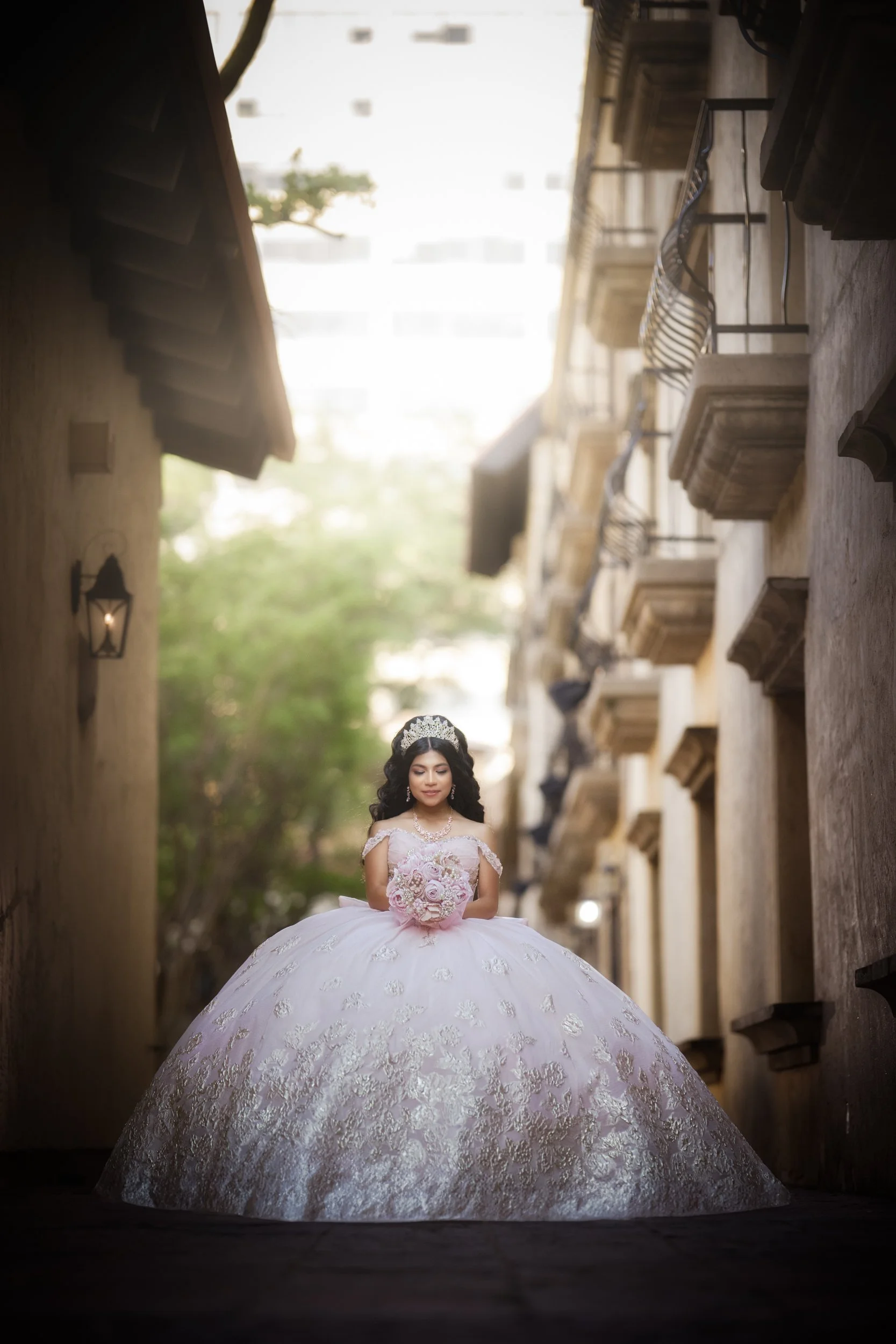 Centered between historic stone buildings, the quinceañera is framed symmetrically in a full-length portrait. Her voluminous gown spreads beautifully around her as she looks down at her bouquet, evoking a serene, regal moment enhanced by natural ligh