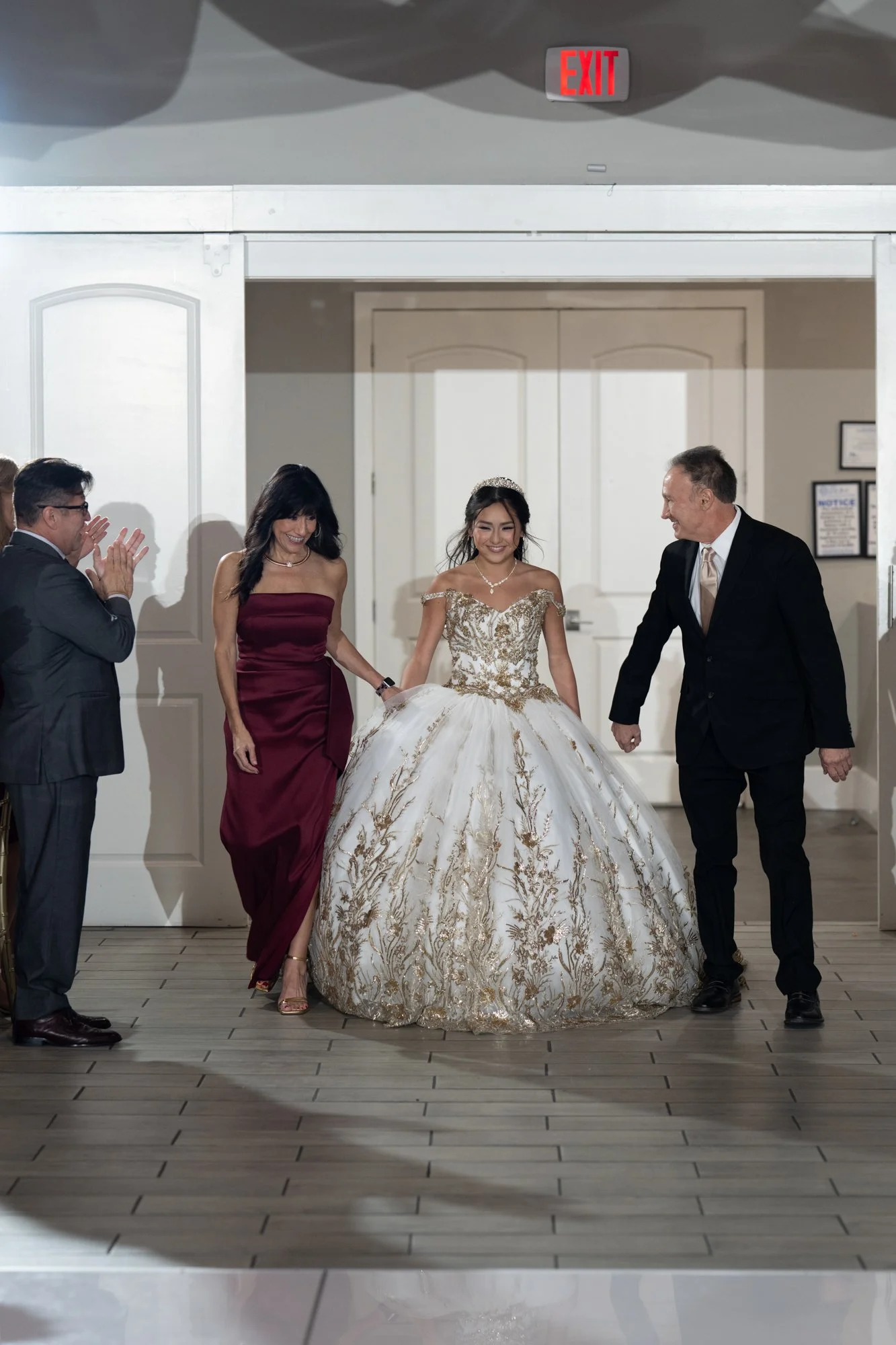 A powerful entrance moment as Alex is escorted into her reception by her parents. Dressed in her elegant white and gold embroidered ball gown, she smiles with confidence and grace while guests applaud her arrival. The moment perfectly captures the pr