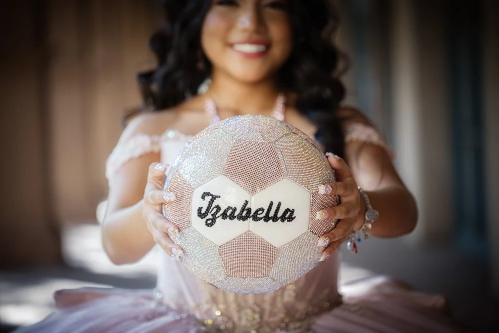 A beautiful Quinceañera proudly presents a custom rhinestone soccer ball featuring her name, blending her passion for sports with the elegance of her milestone celebration. This creative portrait captures her personality, achievements, and unique jou