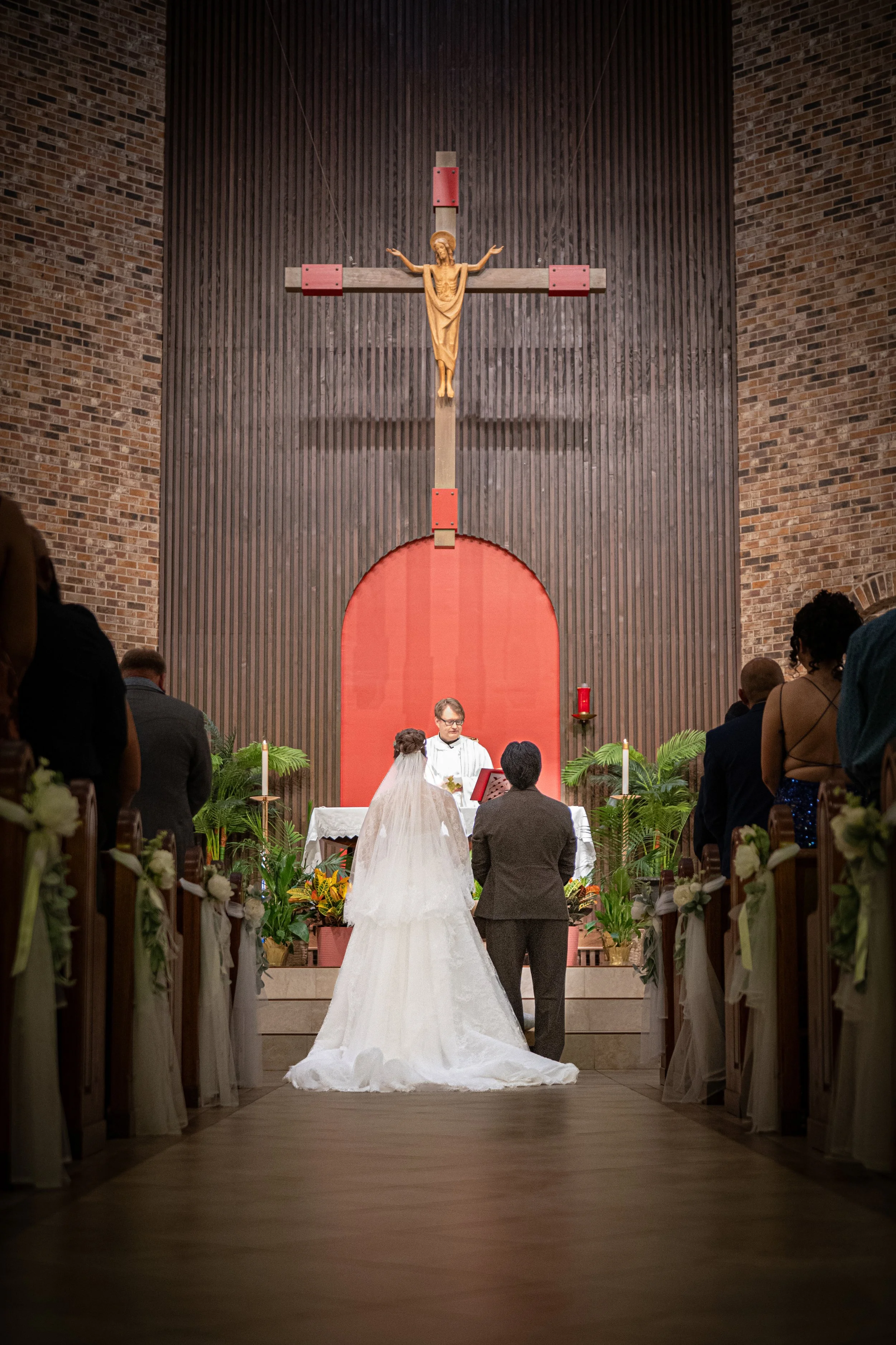 Bride and groom standing at the altar during a Catholic wedding ceremony, framed by the church aisle with guests seated on both sides as they exchange vows beneath a large crucifix.