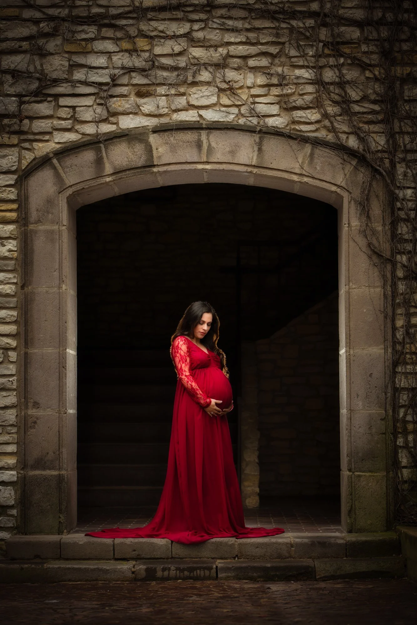 A fine-art maternity portrait of a pregnant woman standing in a stone archway, wearing a deep red lace gown. She gently cradles her belly while soft shadows and textured stone walls frame an intimate and timeless moment.