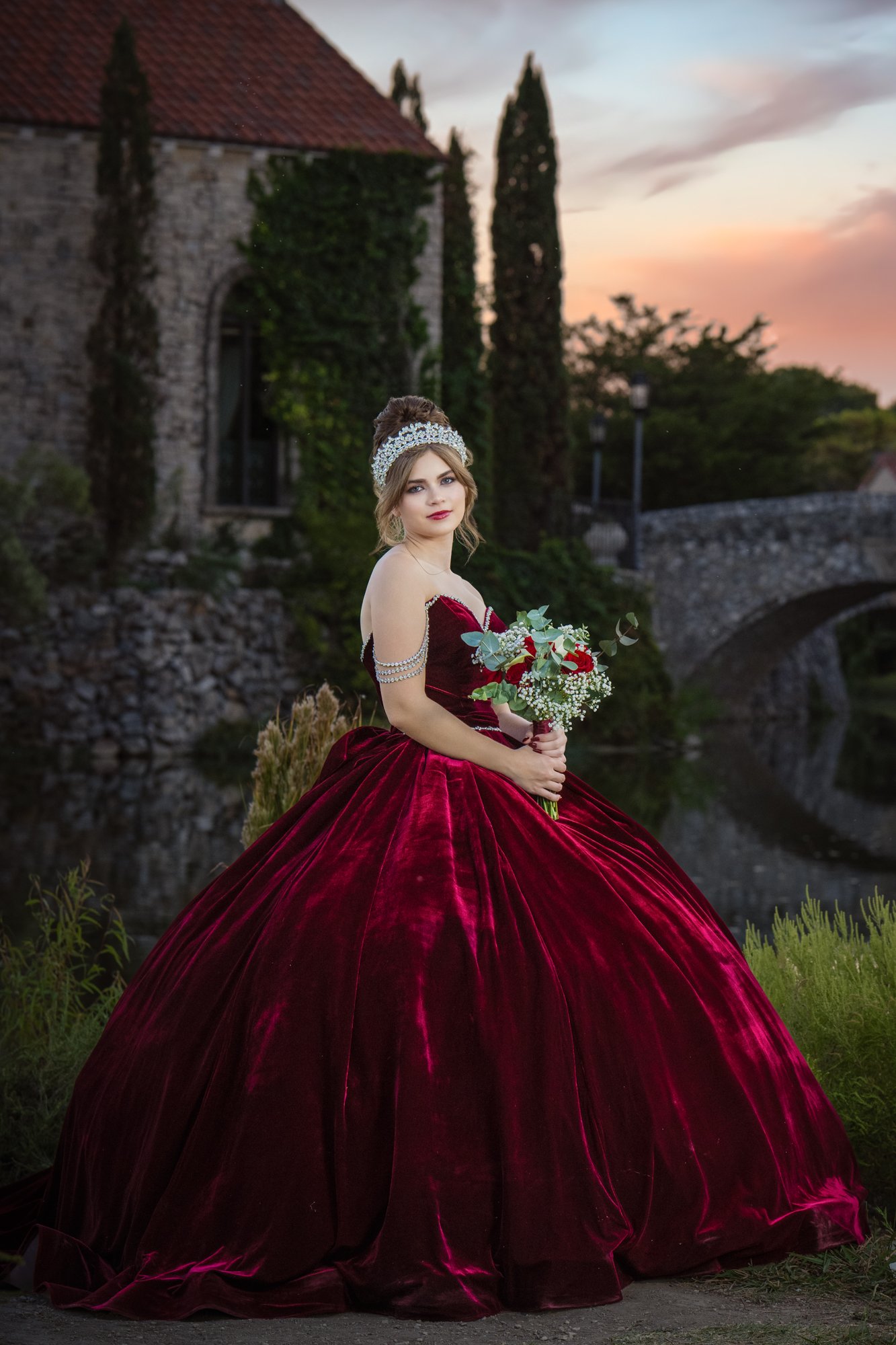 A quinceañera in a deep burgundy velvet ball gown holds a floral bouquet while posing gracefully near a stone bridge at sunset, creating a romantic quinceañera portrait that blends elegance, tradition, and timeless European-inspired scenery.