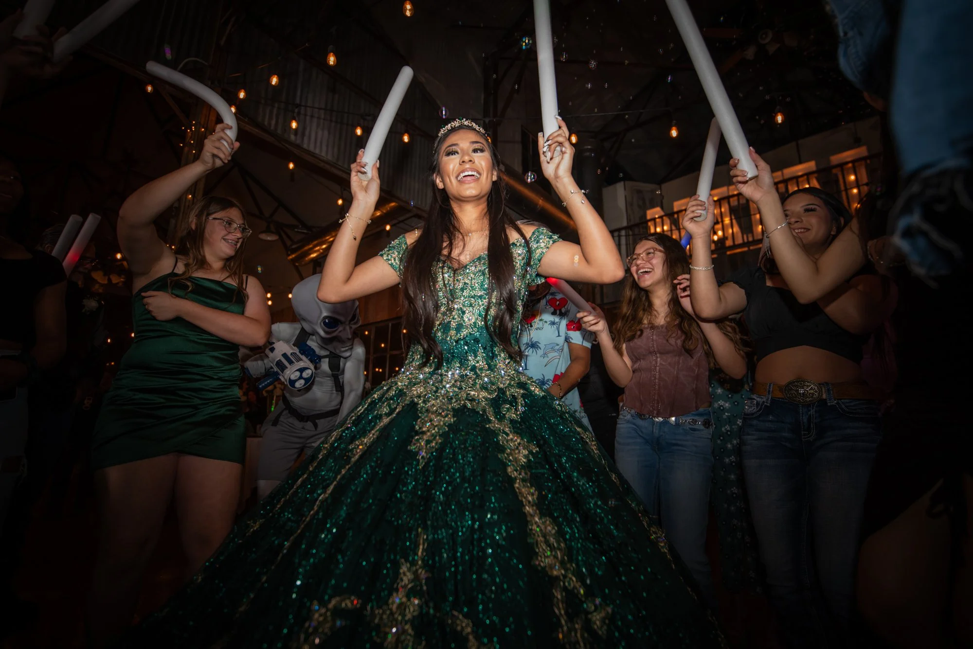 Hands raised, glowing foam sticks in the air, and pure joy on her face, this is the energy of a quinceañera fully embracing her night. The emerald gown sparkles against the warm string lights, making her the undeniable center of the celebration.