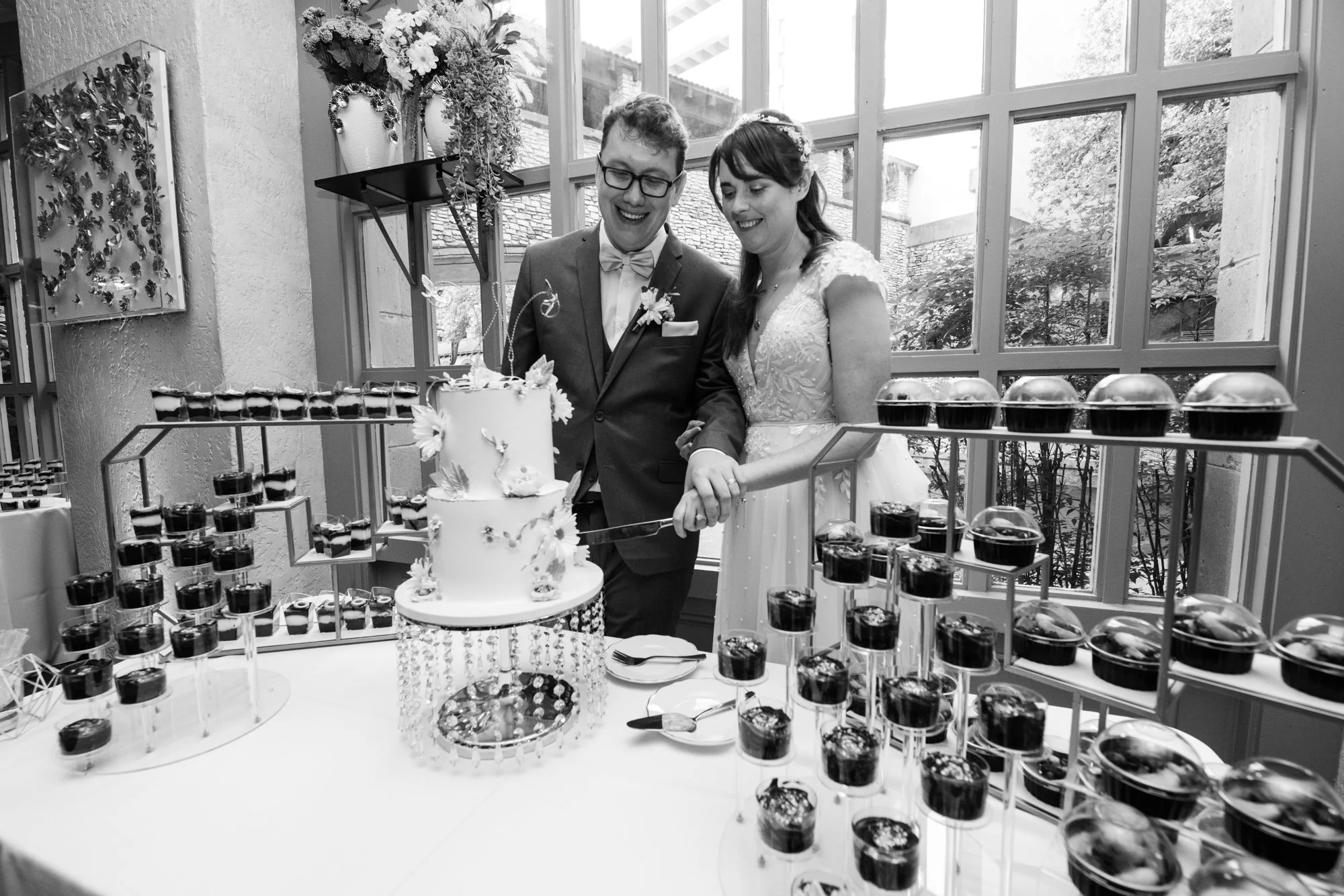 A black-and-white image of the bride and groom cutting their wedding cake together indoors, surrounded by elegant dessert displays and natural window light, highlighting an intimate reception moment.