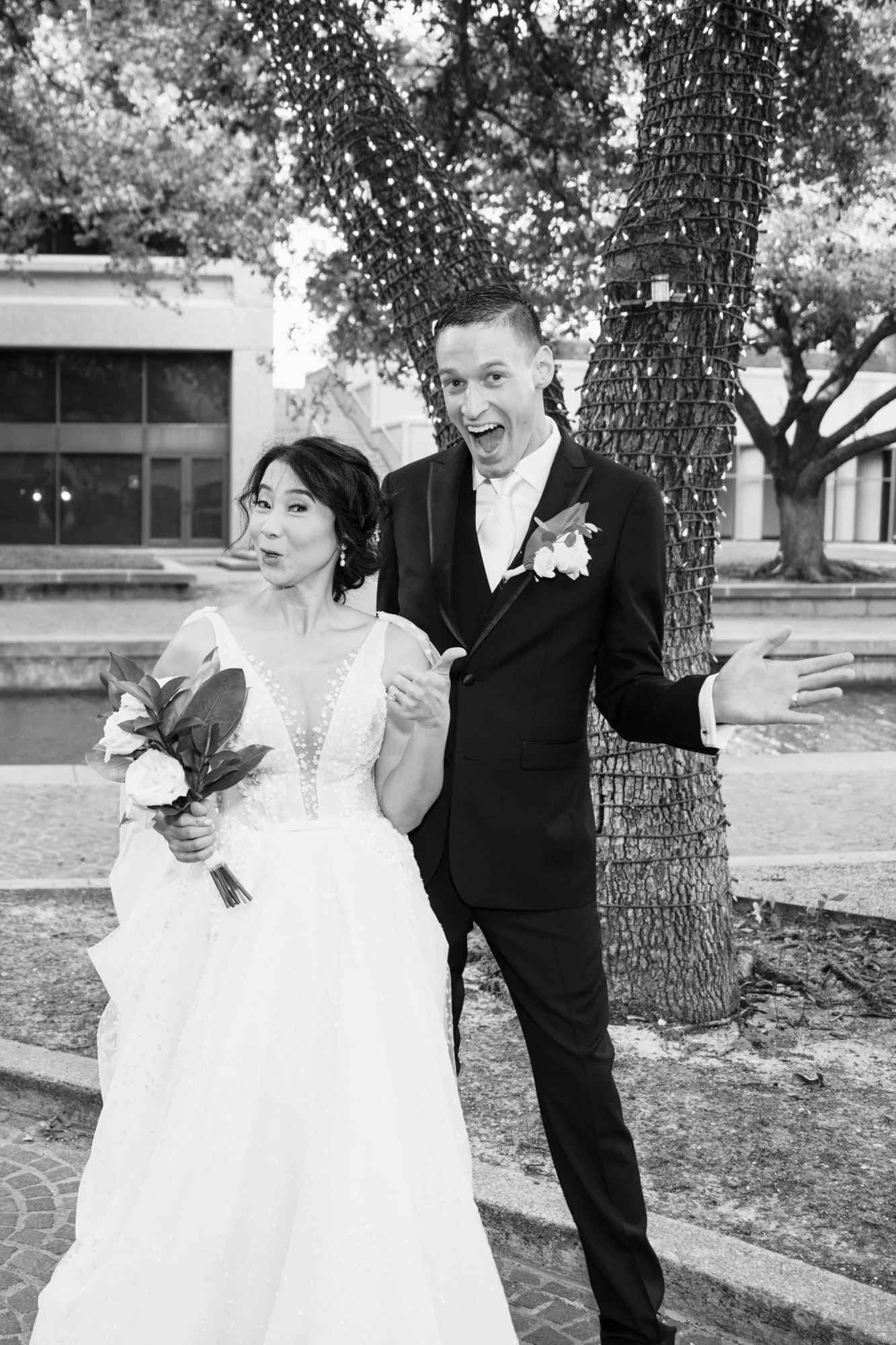 A playful black-and-white portrait of the bride and groom standing together outdoors beneath tree lights. The bride holds her bouquet while both react with expressive, joyful gestures, showcasing their personalities and lighthearted connection.