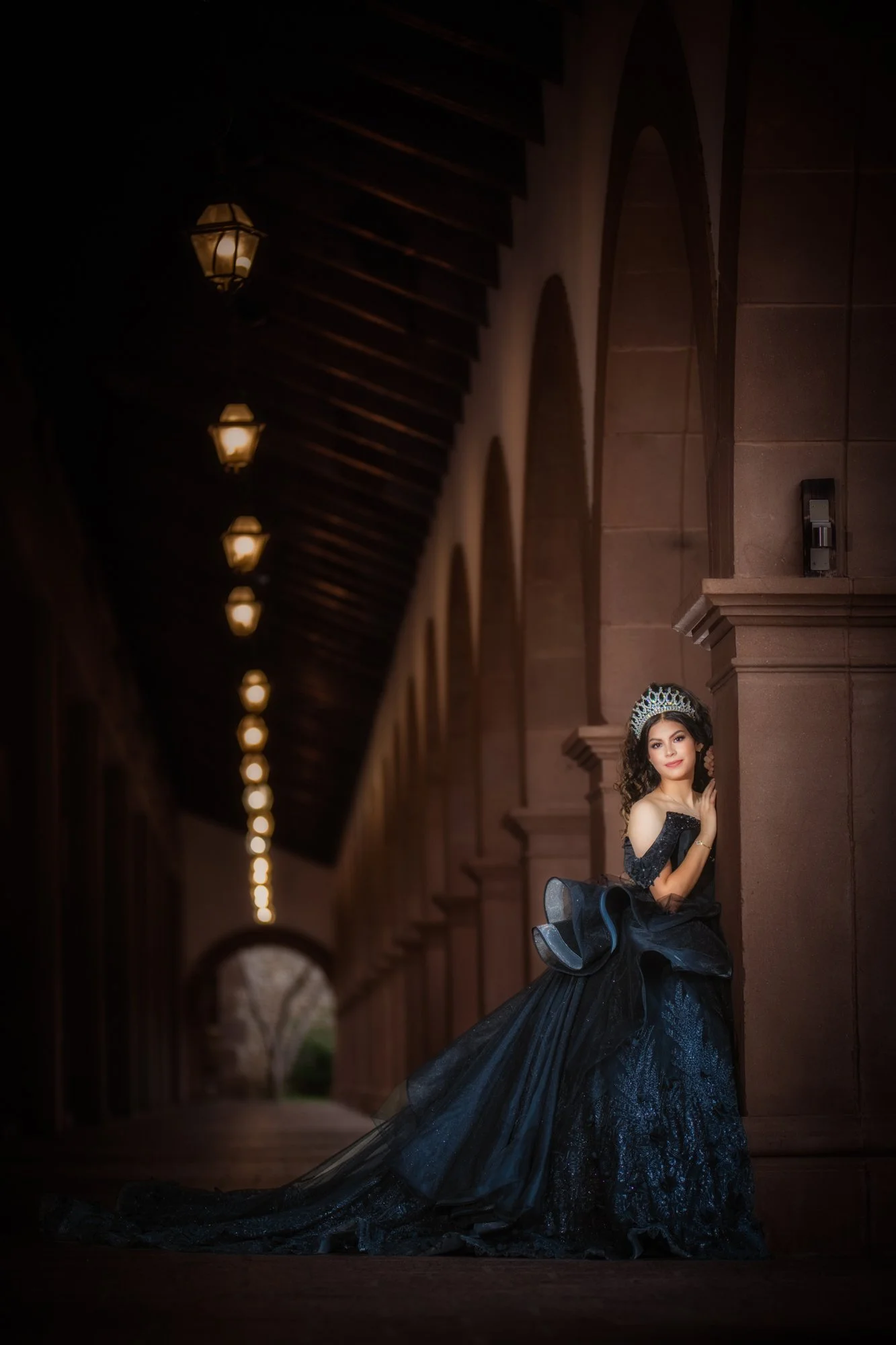 An intimate side-pose moment where the quinceañera gently leans against a stone column, her gown flowing behind her. The soft lighting and architectural lines create a timeless, editorial-style portrait.