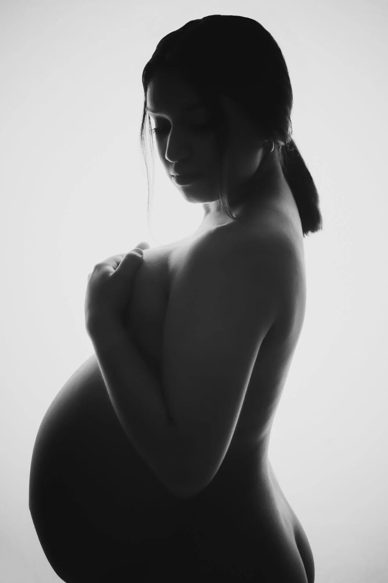 A high-contrast black-and-white maternity silhouette portrait of an expecting mother in profile. Soft studio lighting highlights the curve of her baby bump and facial features, emphasizing strength, serenity, and maternal beauty.