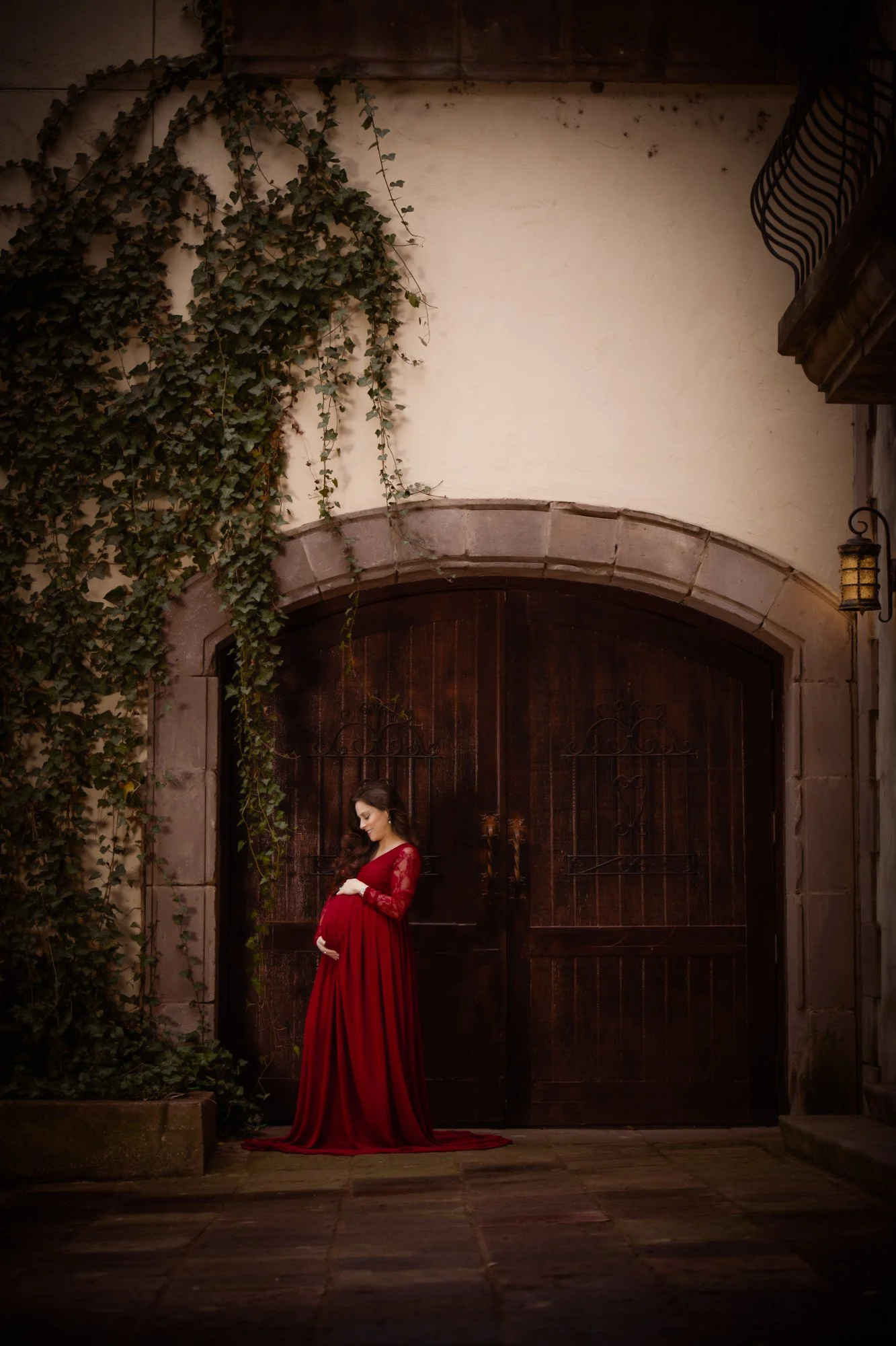 A cinematic maternity portrait of an expecting mother in a flowing red gown posed in front of large wooden doors covered with ivy. The contrast between the rich red fabric and the aged stone architecture creates a romantic, storybook atmosphere