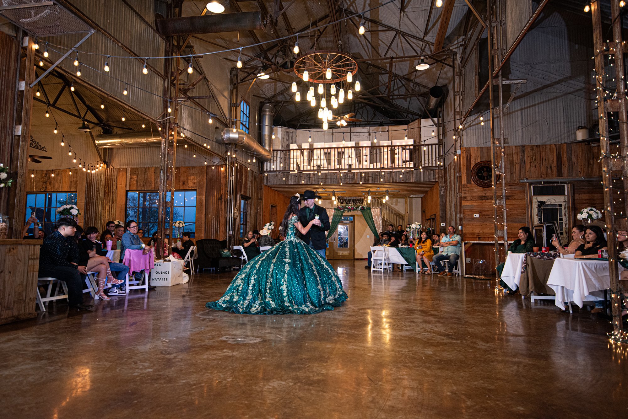 Centered in a rustic industrial venue filled with chandeliers and warm ambient lighting, Natalie glides across the dance floor during her traditional father, daughter dance. The sweeping emerald skirt reflects softly against the polished concrete, cr