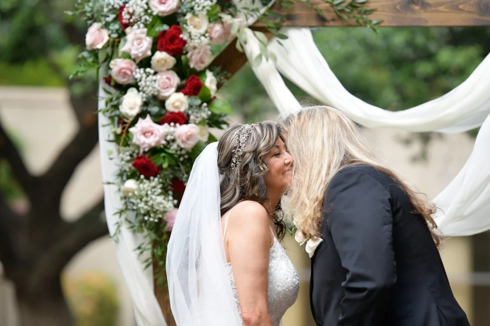Bride and groom sharing a kiss during their outdoor wedding ceremony beneath a floral arch decorated with red, blush, and white roses.