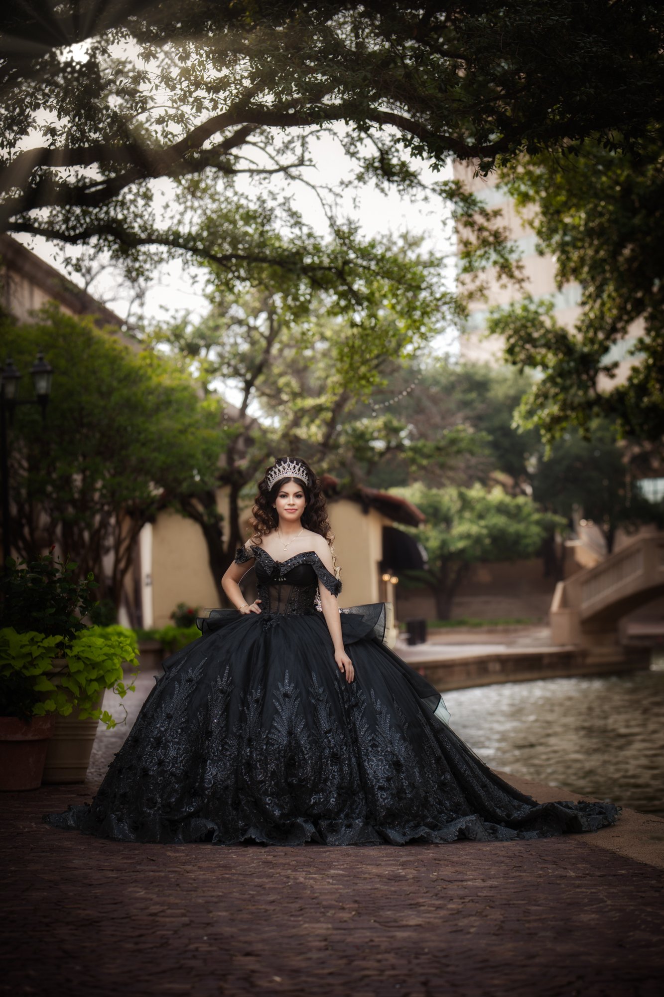 The quinceañera stands confidently beneath tree canopies along the canal, her black gown cascading gracefully as soft natural light filters through the leaves. This portrait highlights confidence, maturity, and the beauty of the moment.