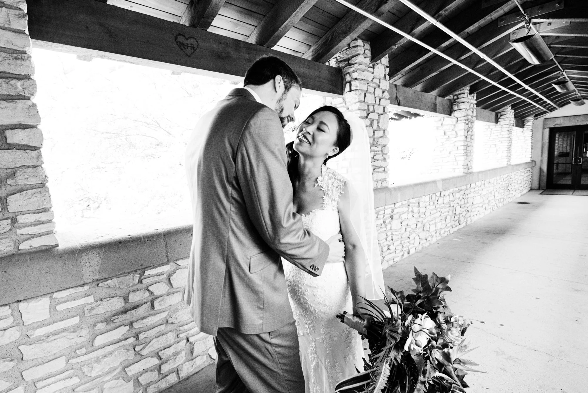A quiet, candid portrait of the bride and groom sharing a laugh beneath a covered stone walkway. The bride holds her bouquet close as they lean into one another, radiating warmth and connection.