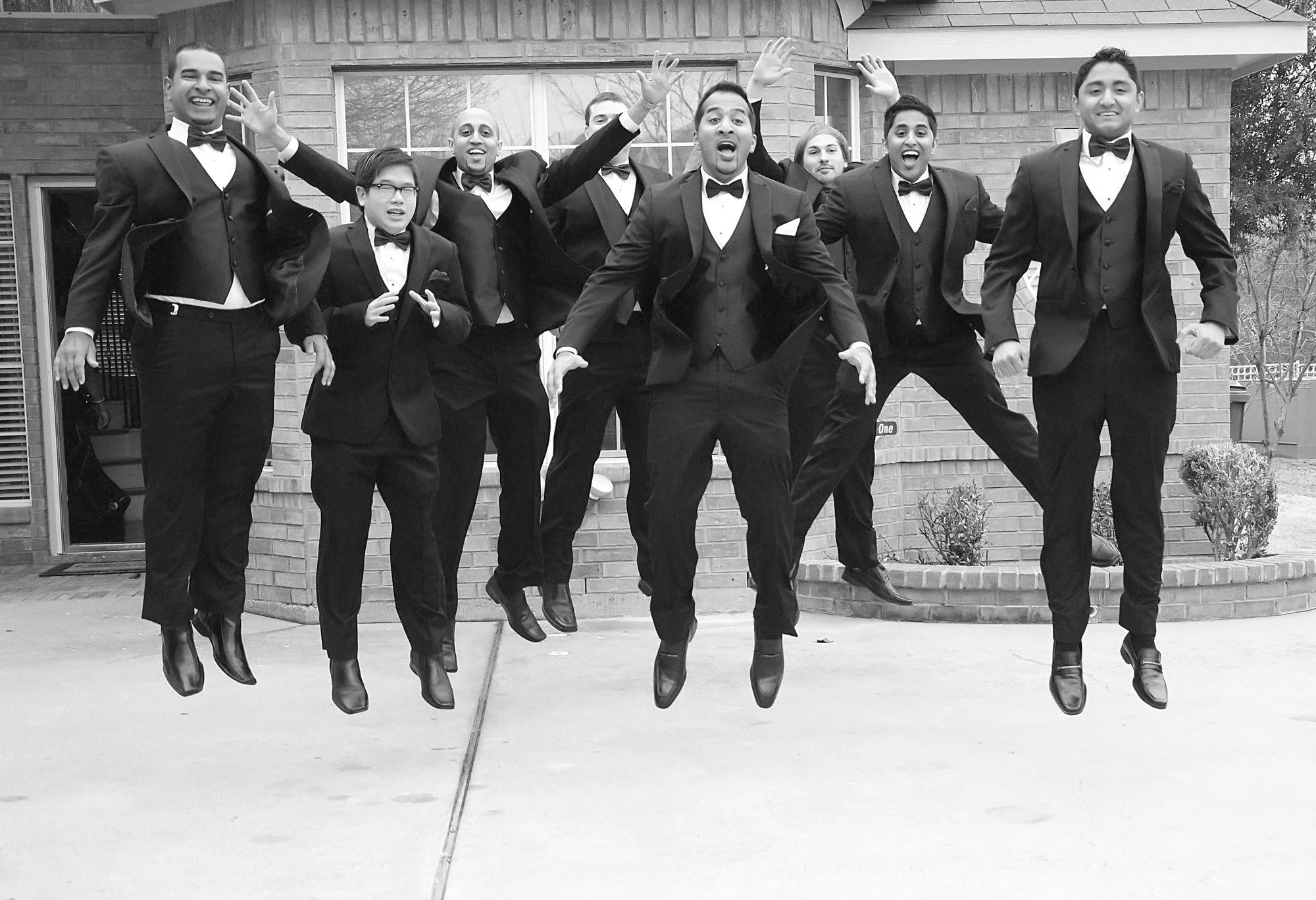 The groomsmen leap into the air together in unison, captured mid-jump in a high-energy black-and-white portrait. Their expressions and movement convey excitement, friendship, and celebration.
