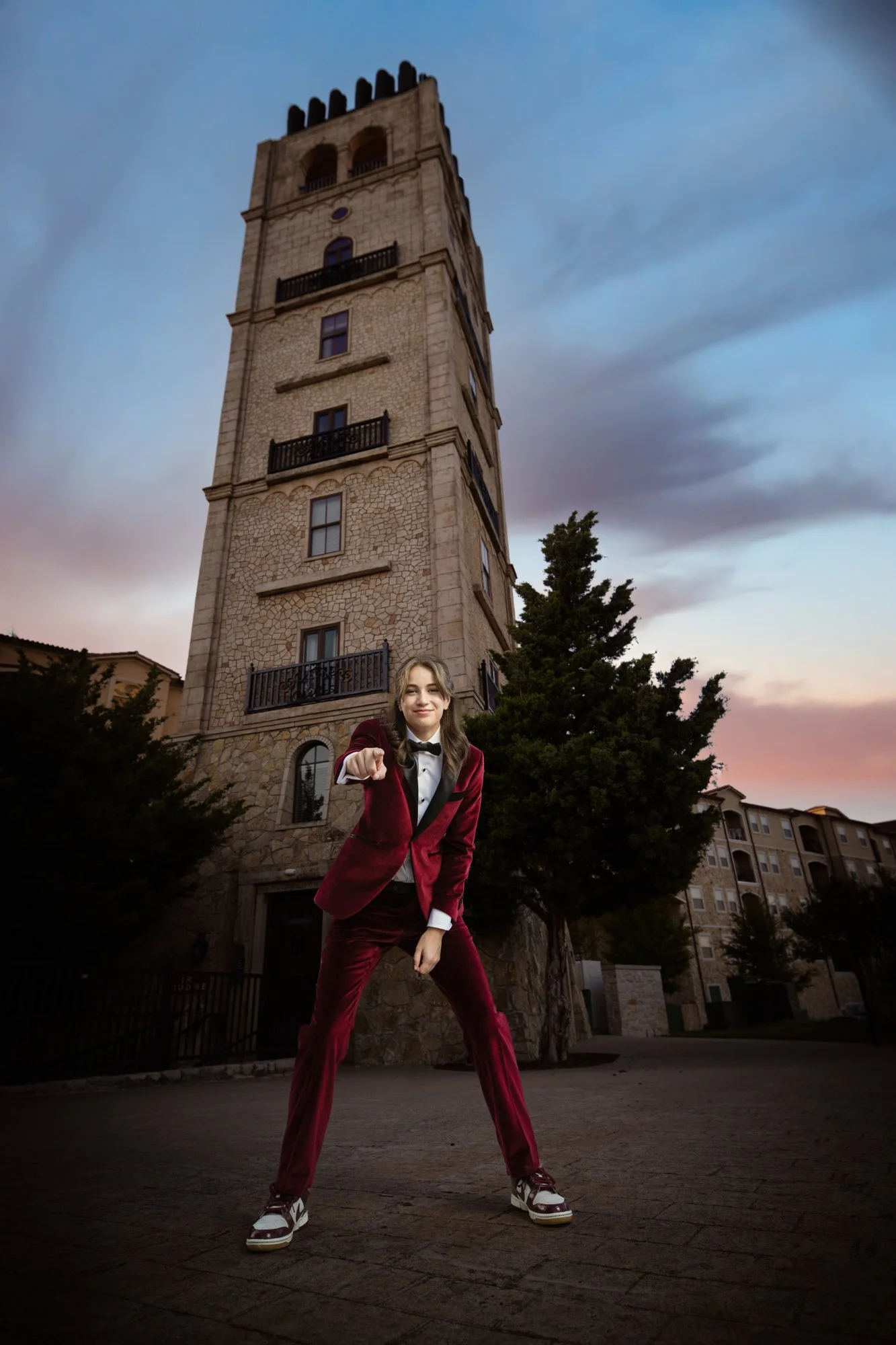 A stylish quinceañera in a burgundy tuxedo points playfully toward the camera in front of a historic stone tower at sunset, capturing personality, confidence, and modern flair within a classic quinceañera portrait setting.
