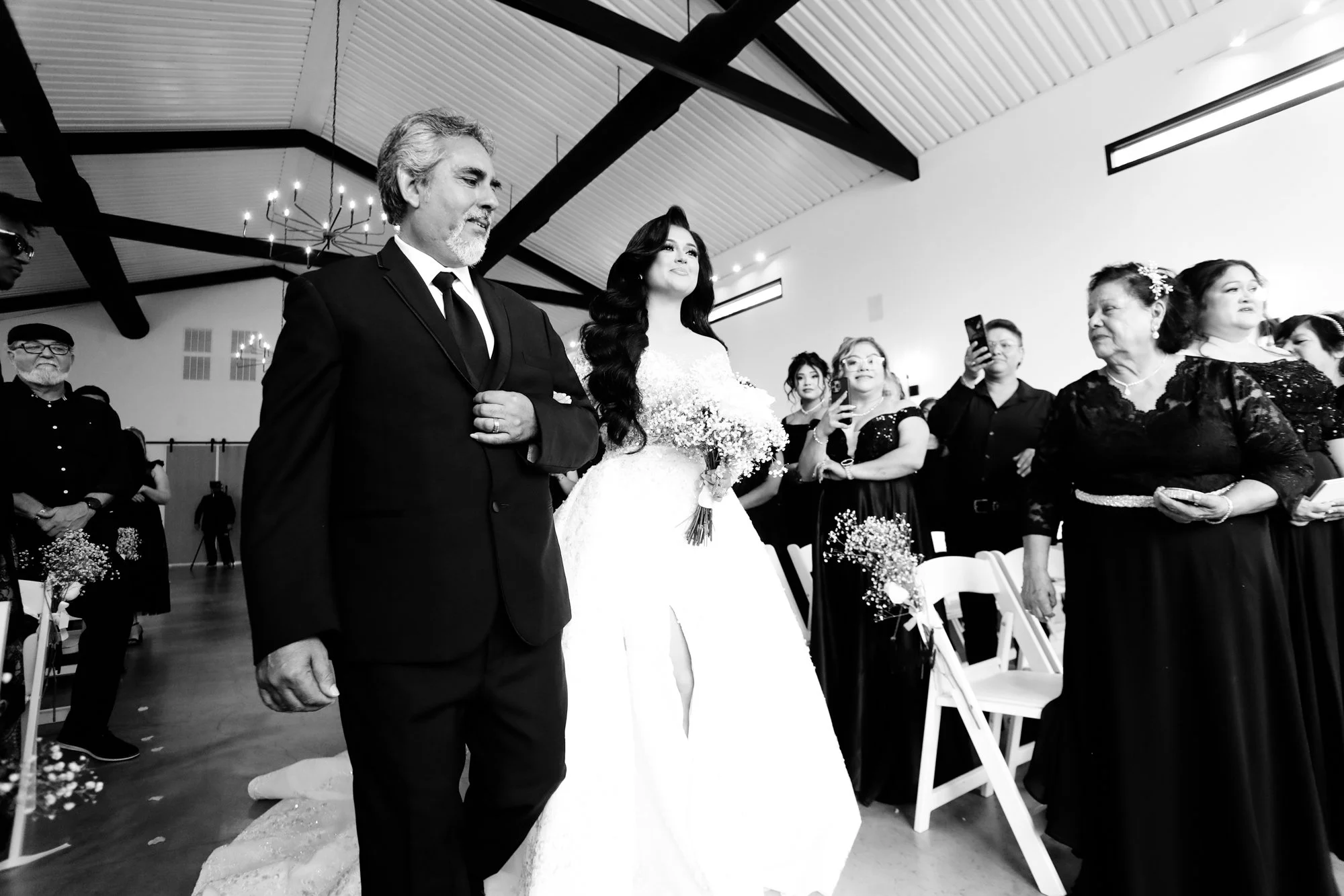 Bride walking down the aisle arm-in-arm with her father during an emotional wedding ceremony, surrounded by guests capturing the moment in a bright, airy chapel.