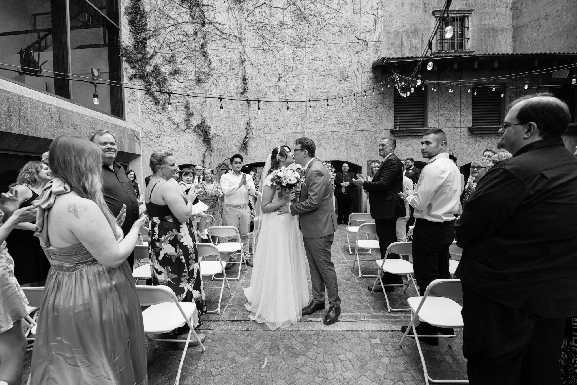 A wide black-and-white ceremony shot of the bride and groom sharing their first kiss as guests stand and applaud on both sides of the aisle in an outdoor courtyard venue.