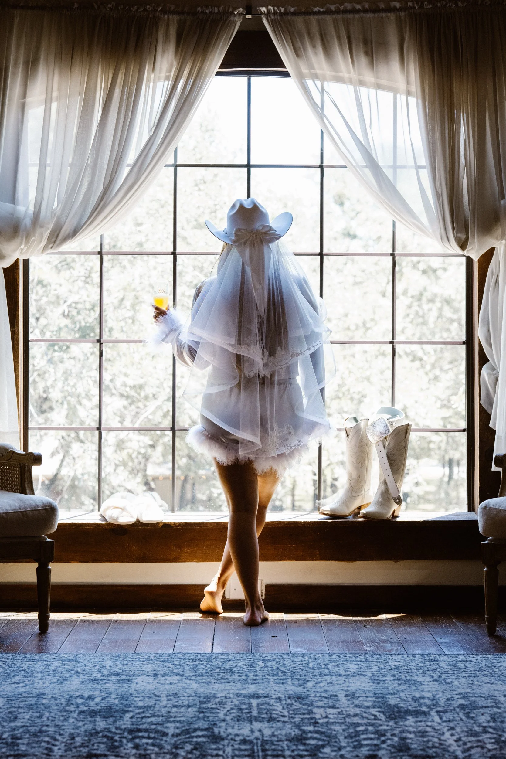 A striking silhouette of the bride standing barefoot at a window in her robe and veil, blending softness and strength in an artistic, storytelling image that defines quiet anticipation.