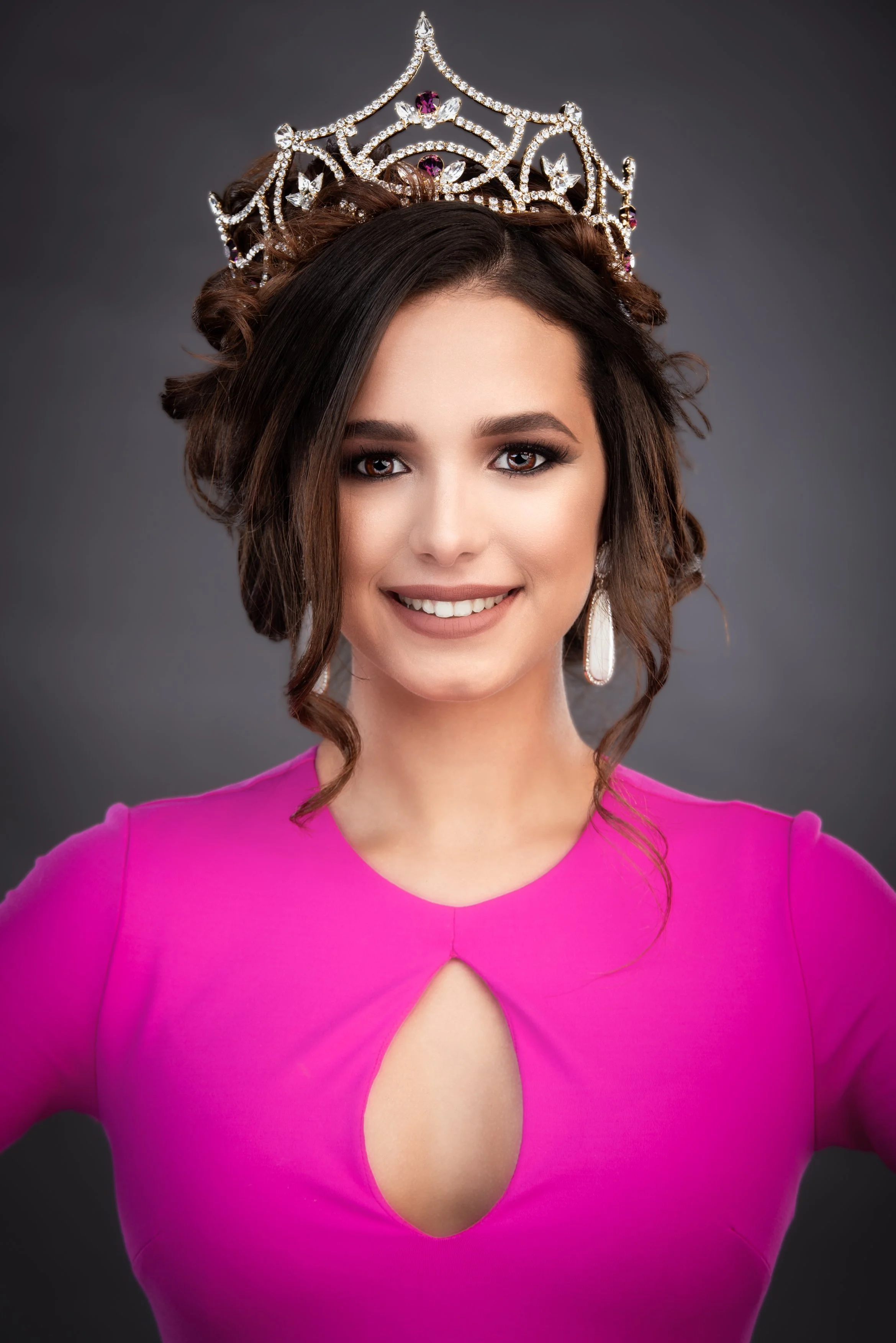 A studio portrait of a young woman wearing an ornate jeweled crown and a vibrant magenta dress with a keyhole neckline. She faces the camera with a confident, warm smile, showcasing polished makeup, soft curled hair styled into an elegant updo, and s