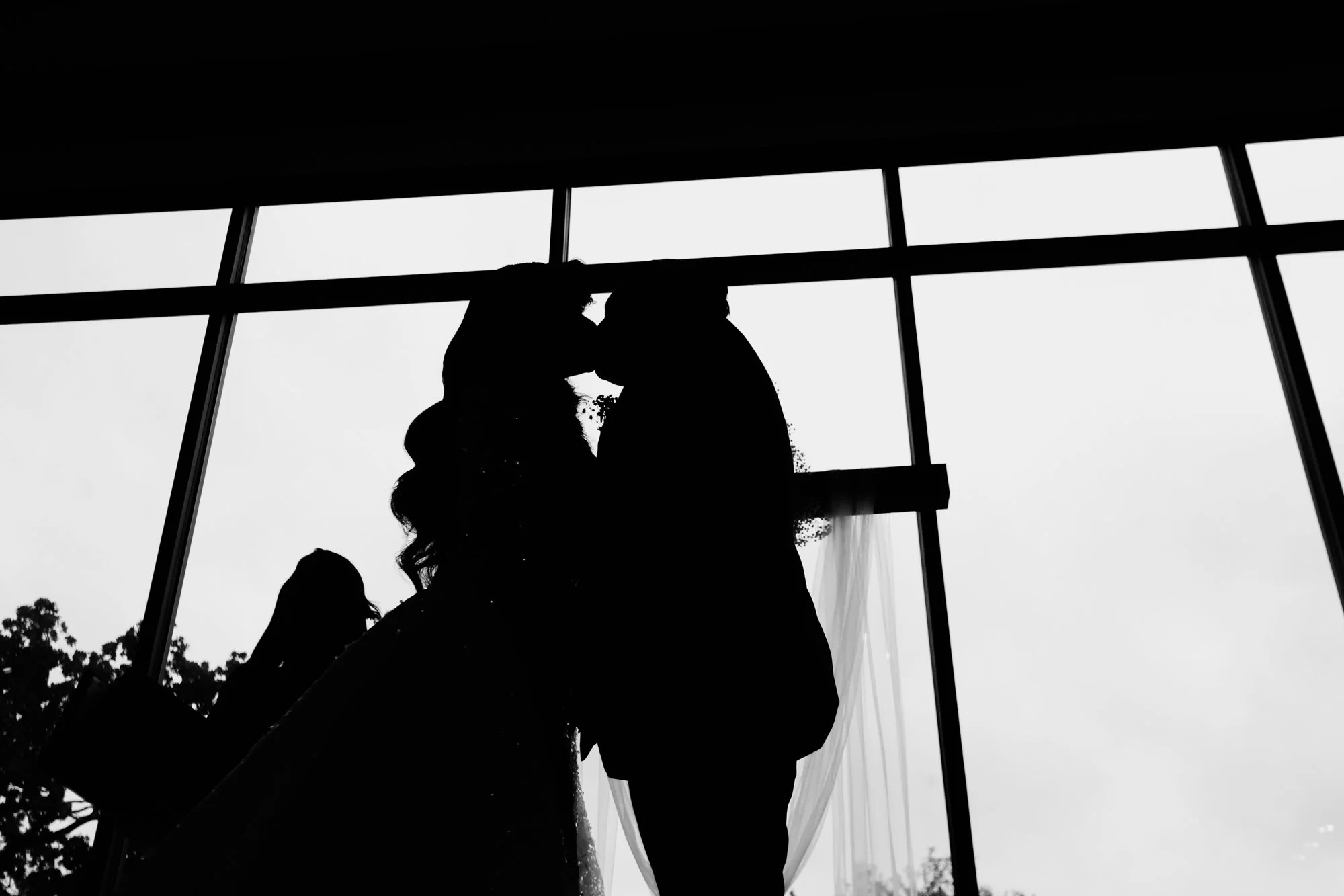 Silhouette of a bride and groom sharing a romantic kiss in front of floor-to-ceiling windows during their wedding reception, captured by a Dallas-Fort Worth wedding photographer.