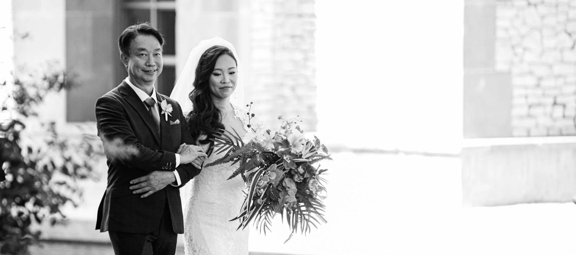 A black-and-white photograph of a bride standing beside her father, holding a cascading floral bouquet, as they pause together before the ceremony in a softly lit courtyard setting.