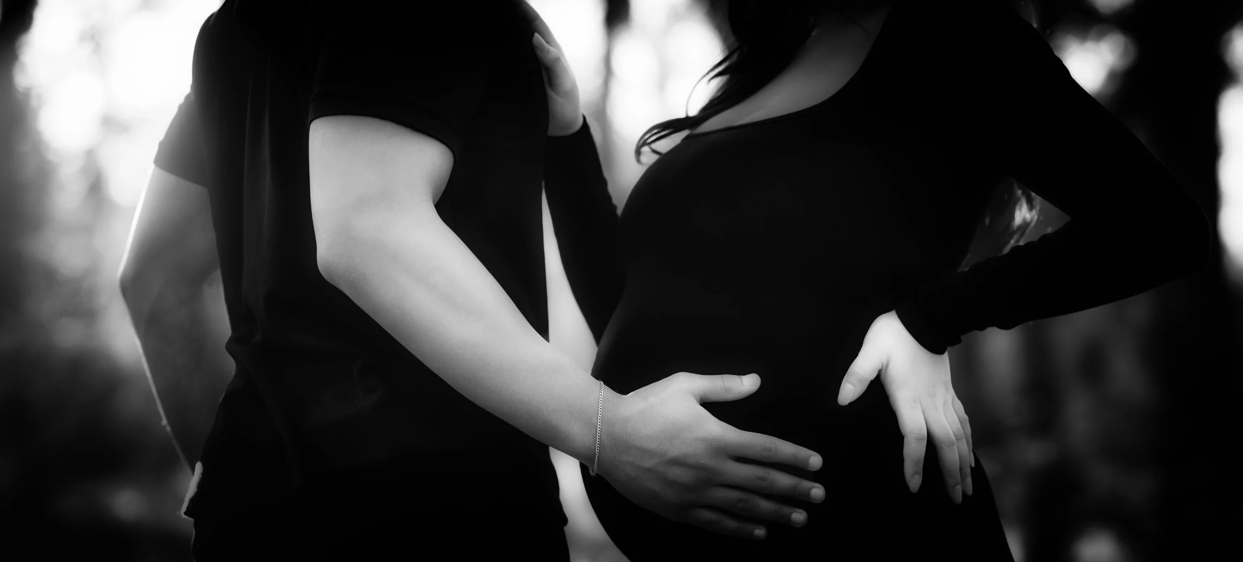 A black-and-white close-up captures an intimate moment as a partner’s hand gently rests on a pregnant belly, symbolizing connection, protection, and anticipation of new life. The shallow depth of field adds softness and emotional depth.