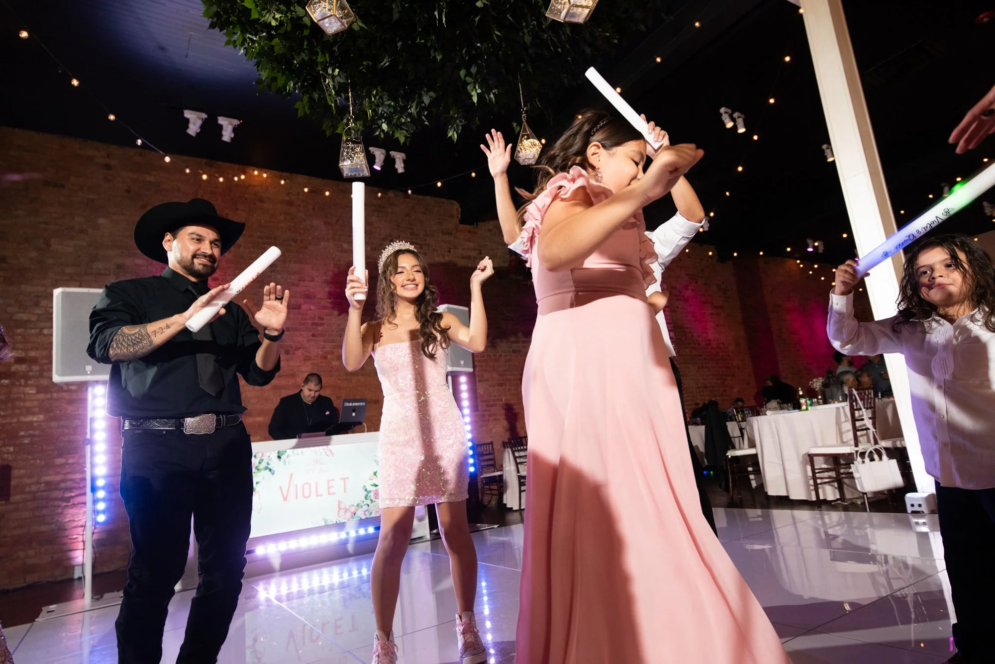 The energy shifts to full celebration mode. Surrounded by family and friends waving glow sticks, the quinceañera beams with excitement on the dance floor, balancing tradition with vibrant party vibes.