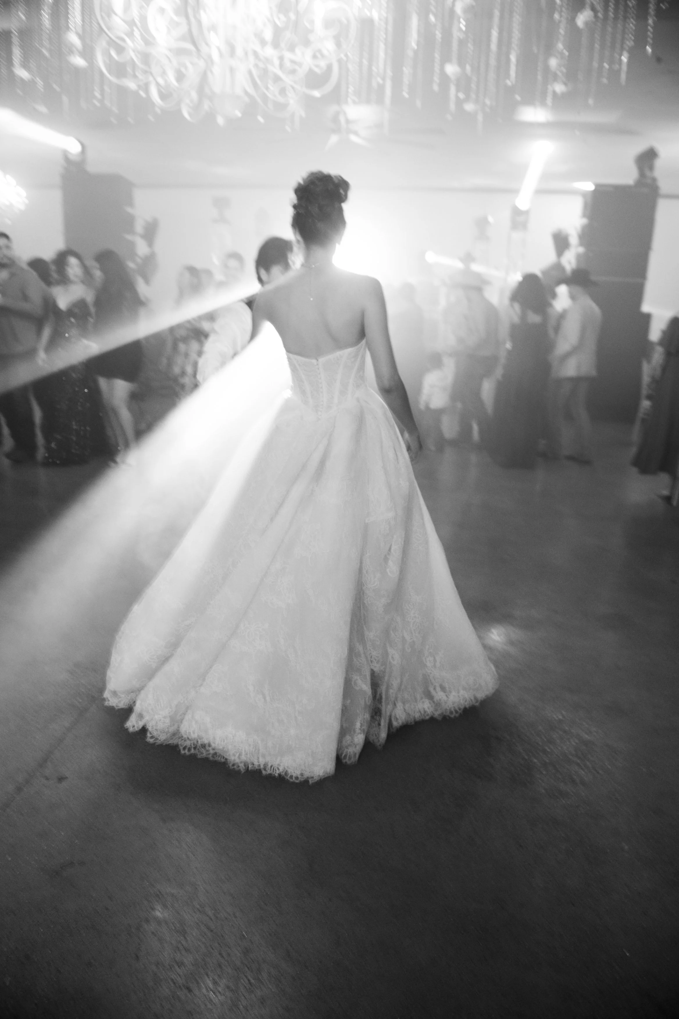 A black-and-white image of the bride walking alone across the dance floor from behind, her gown flowing as stage lighting and guests create a powerful, spotlighted silhouette.