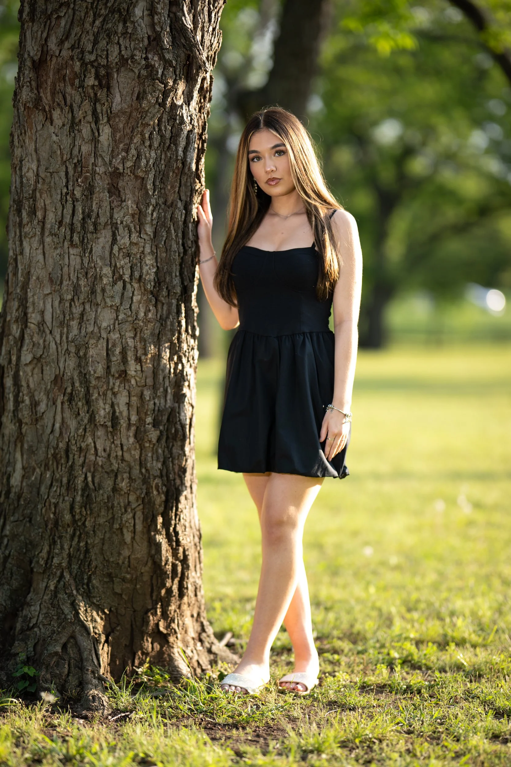 A young woman in a black dress stands beside a large tree in a sunlit park, gazing calmly toward the camera with soft golden-hour light filtering through the greenery.