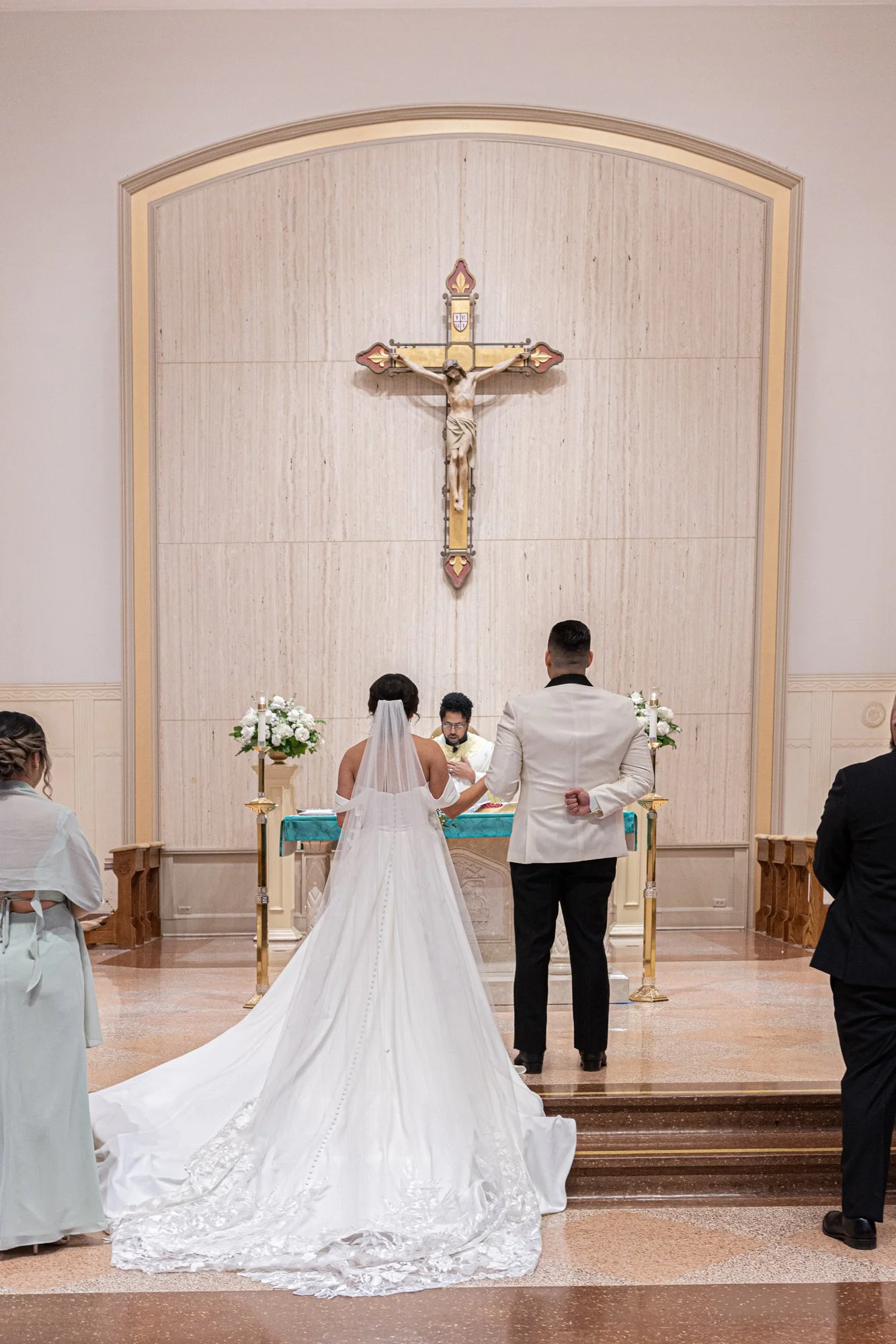 Briseida and Sergio stand before the altar during their Catholic wedding ceremony in Dallas, Texas, exchanging vows beneath a large crucifix as family witnesses their sacred marriage.