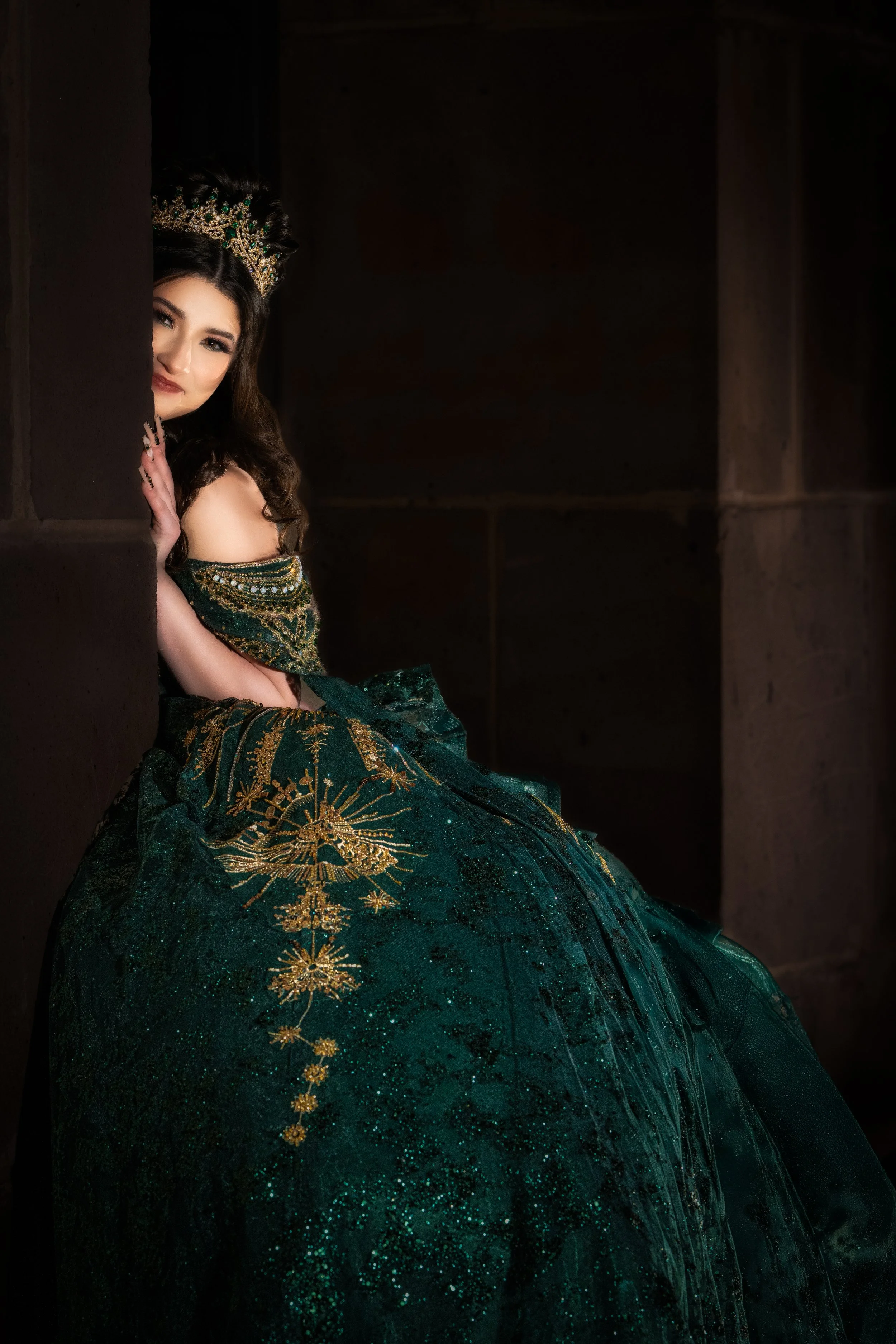 An intimate Quinceañera portrait showing the Quinceañera peeking from behind a stone column, wearing a jeweled crown and emerald gown, blending softness and strength within historic architecture.