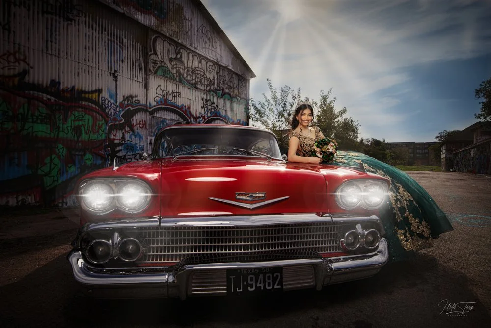 A beautiful Quinceañera poses beside a vintage red classic car, blending elegance and personality in a unique urban portrait session. The dramatic sky and colorful backdrop add a bold and memorable touch to the image.