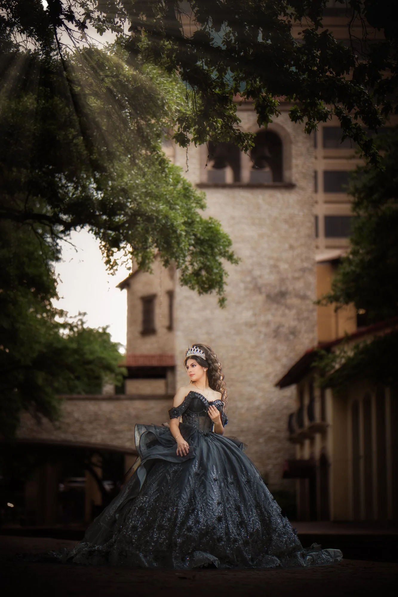 Sunlight streams through the trees, illuminating the quinceañera as she stands poised in her black gown. The historic stone building in the background adds texture and contrast, creating a refined and story-driven image.