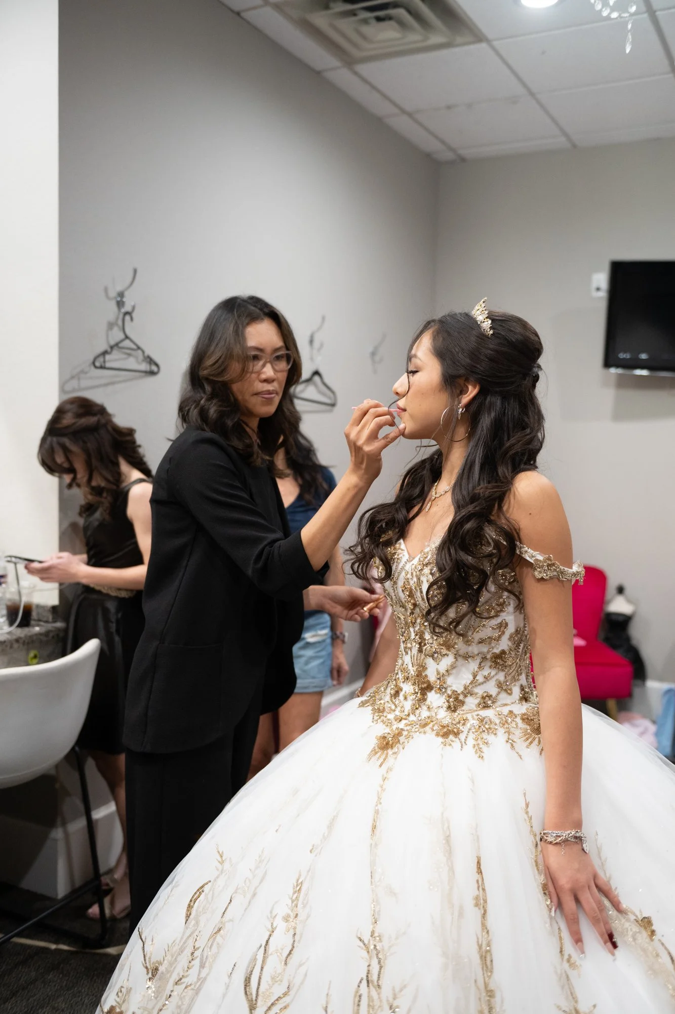 A calm pre-reception moment as Alex receives final makeup touches. Her tiara sparkles under soft lighting, and her gold-detailed gown shines with elegance, capturing the transformation into her full Quinceañera princess look.