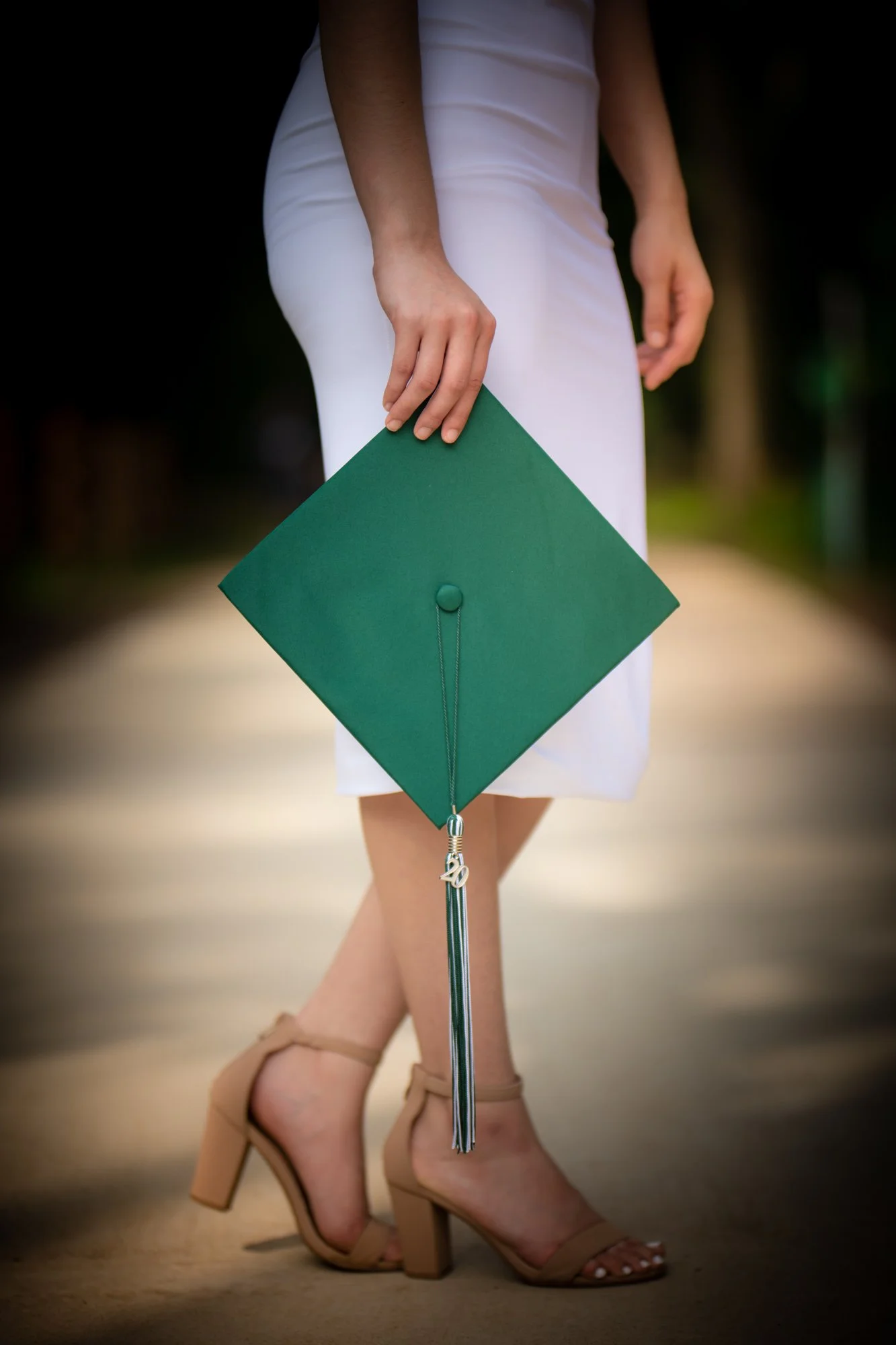 A close-up detail shot of a graduate holding a green mortarboard cap by her side, wearing nude heels and a white dress against a softly blurred outdoor background.
