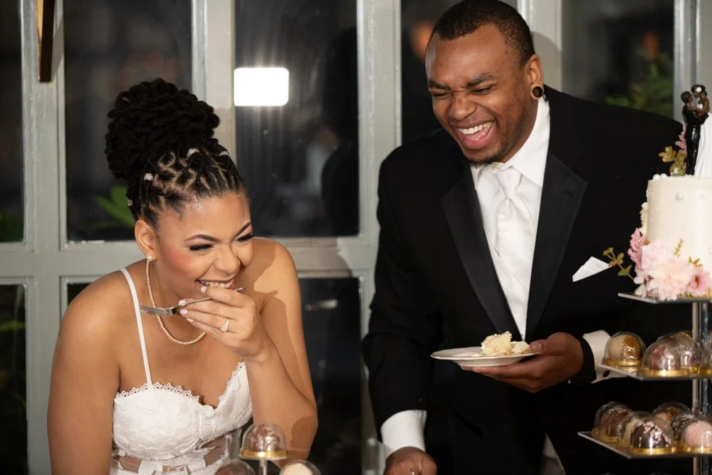 Bride and groom laughing together while enjoying wedding cake and desserts during their reception celebration.