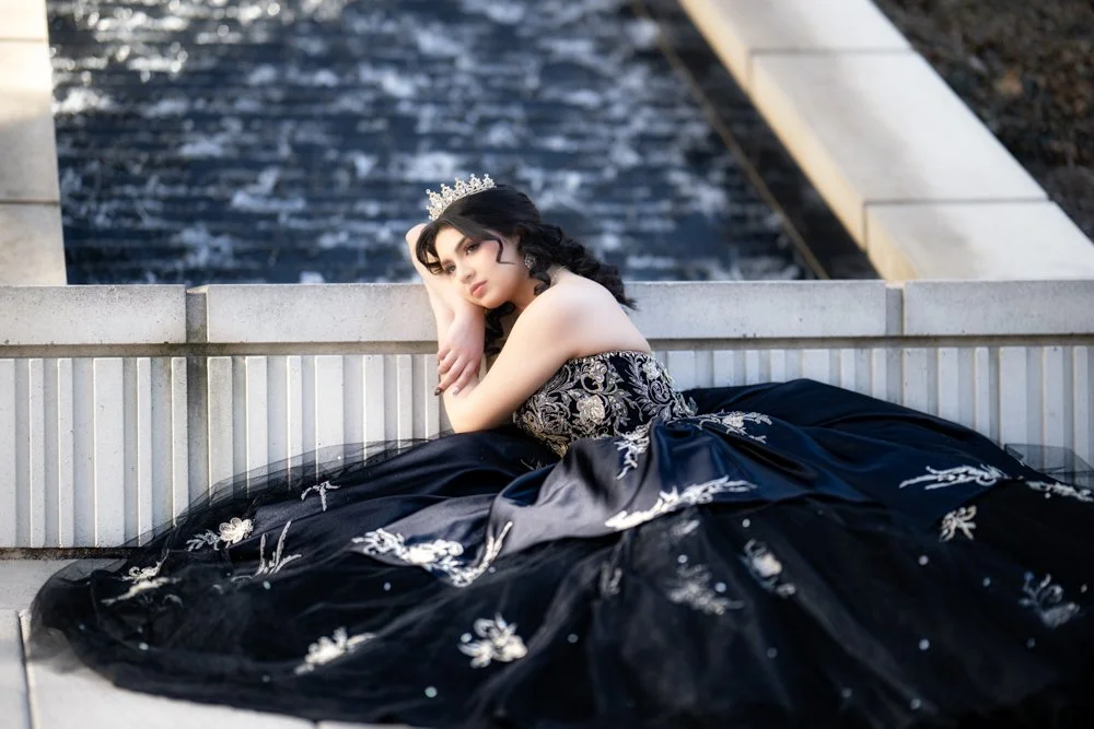 Sofia Moreno relaxes gracefully beside a cascading water feature while wearing a black and gold embroidered Quinceañera gown. The soft lighting and luxurious details create a sophisticated and artistic portrait