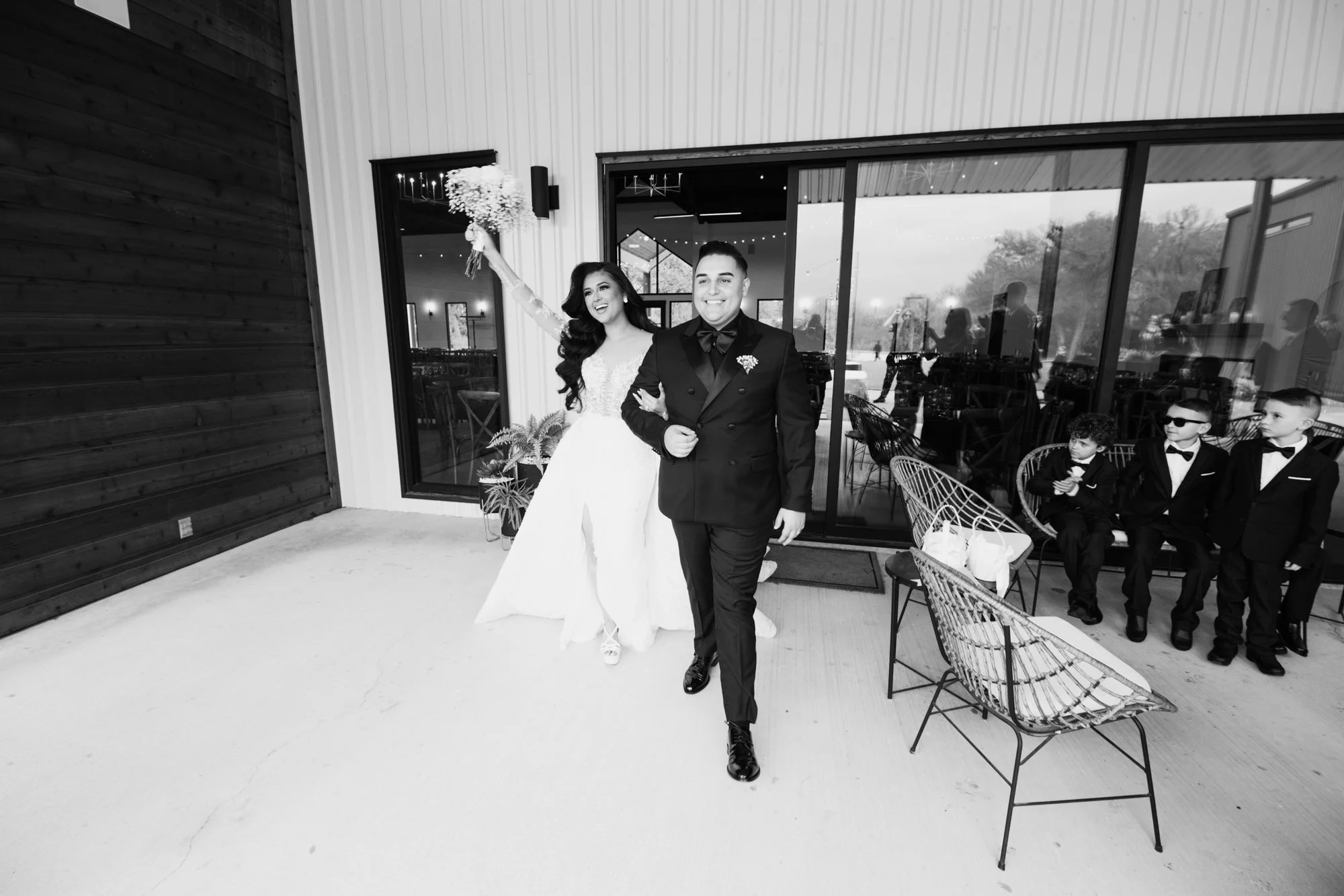 A joyful bride and groom walk hand in hand just after the ceremony, beaming with happiness as the bride raises her bouquet in celebration. Natural light floods the modern venue behind them, capturing the excitement of their first moments as newlyweds
