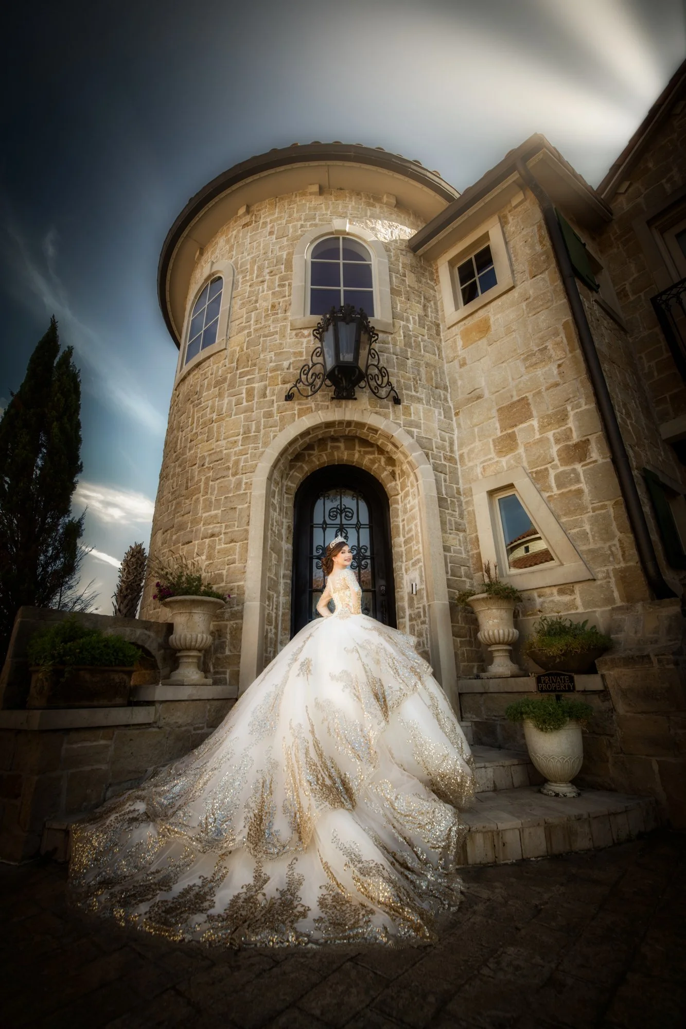 A stunning Quinceañera portrait featuring the young woman posed at the grand entrance of a stone castle, her gold-embroidered ball gown flowing dramatically across the steps. Soft natural light and architectural framing create a regal, fairy-tale atm