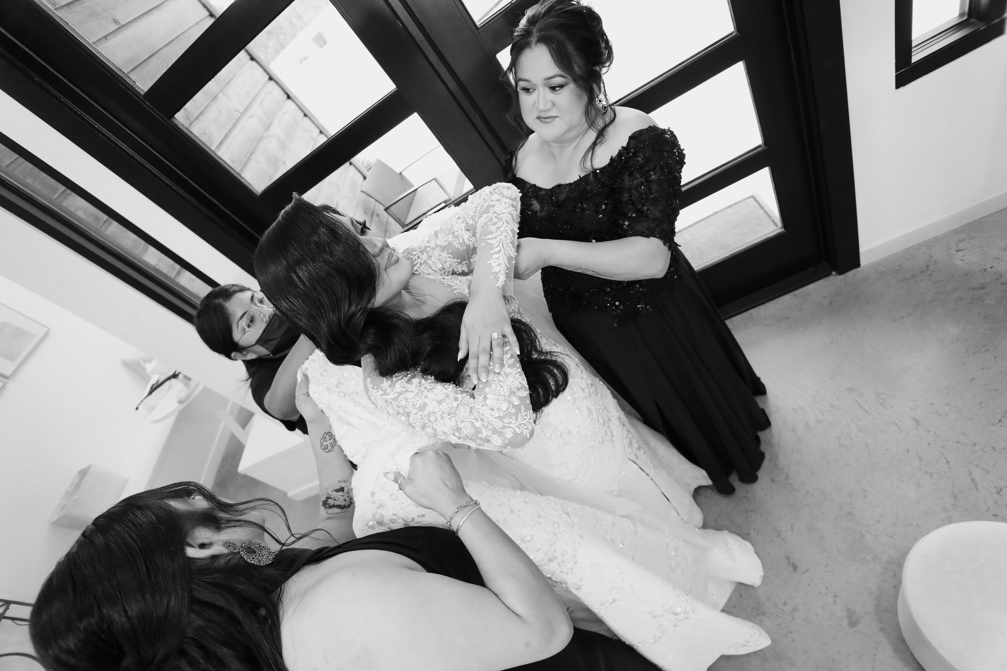 Intimate getting-ready moment as family members help the bride fasten her wedding dress inside a modern bridal suite, highlighting emotion, detail, and connection.