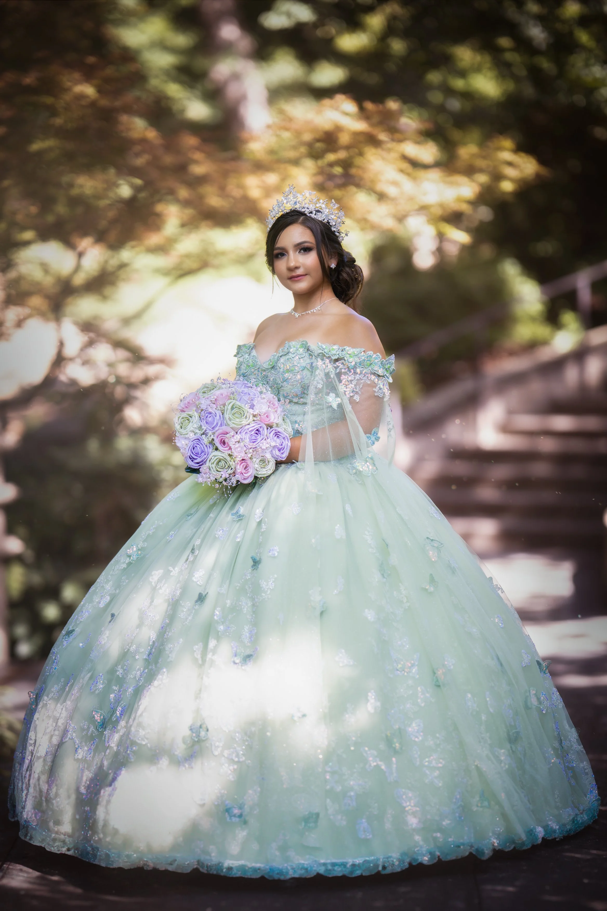 A Quinceañera stands gracefully on a garden path, wearing a mint green ball gown with delicate embroidery, holding a pastel bouquet and wearing a crystal crown, surrounded by soft natural light and blurred trees.