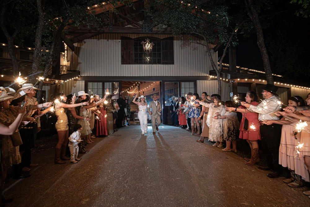Ashley and Jerman walk hand in hand through a sparkler exit surrounded by family and friends outside their beautiful Texas ranch wedding venue at night.