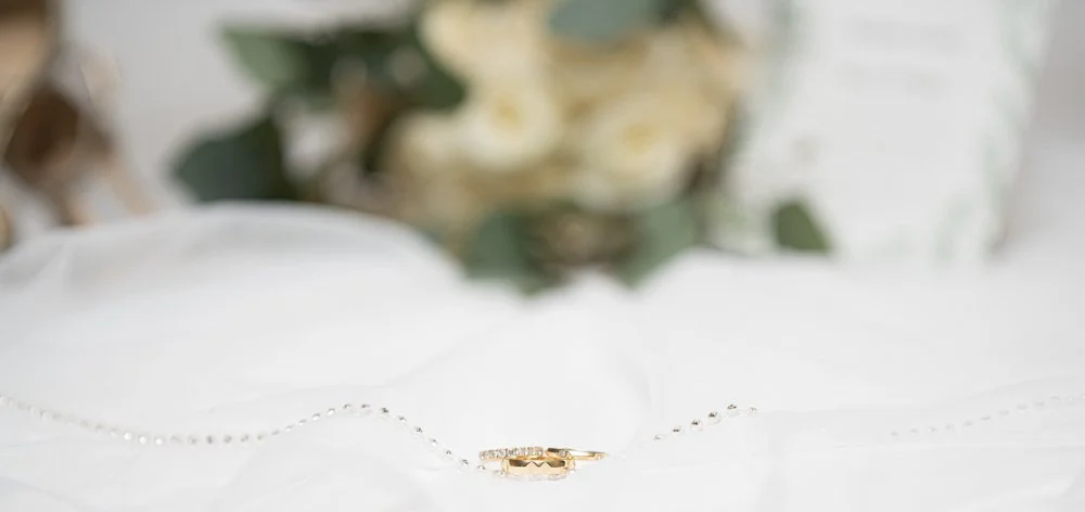 Gold wedding bands displayed on a bridal gown with soft-focus wedding flowers in the background.